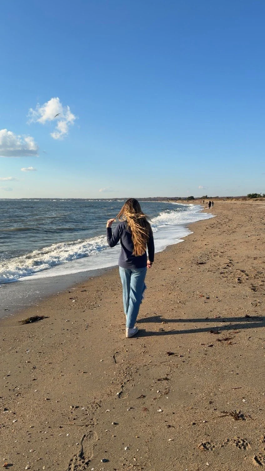 Woman walking along a sandy beach with ocean waves and blue sky, some clouds, and distant figures in the background.
