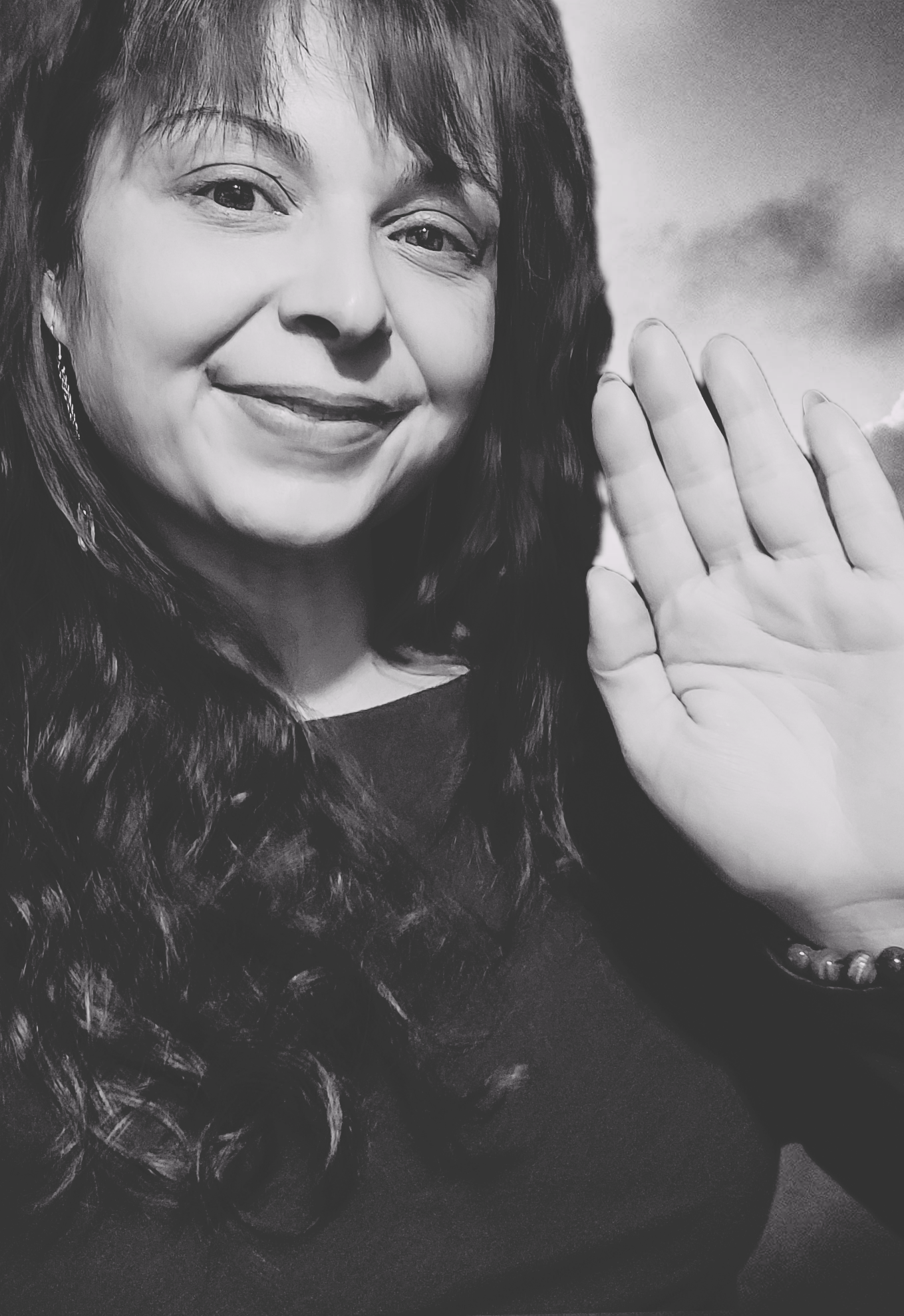 A woman with long dark curly hair smiling and holding up her right hand in a wave gesture.