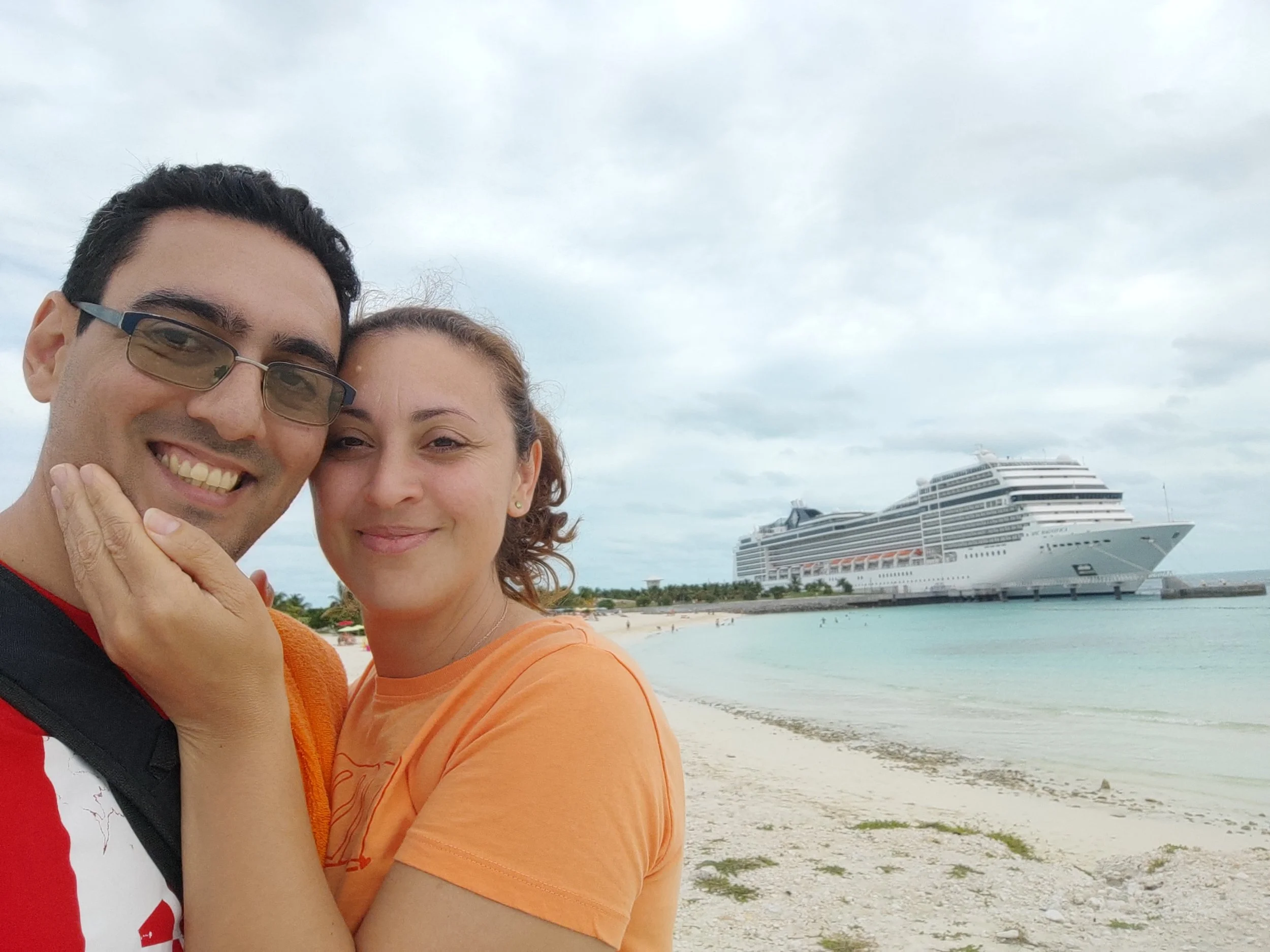 Pareja sonriendo en la playa frente a un crucero de lujo en el mar, con cielo nublado.