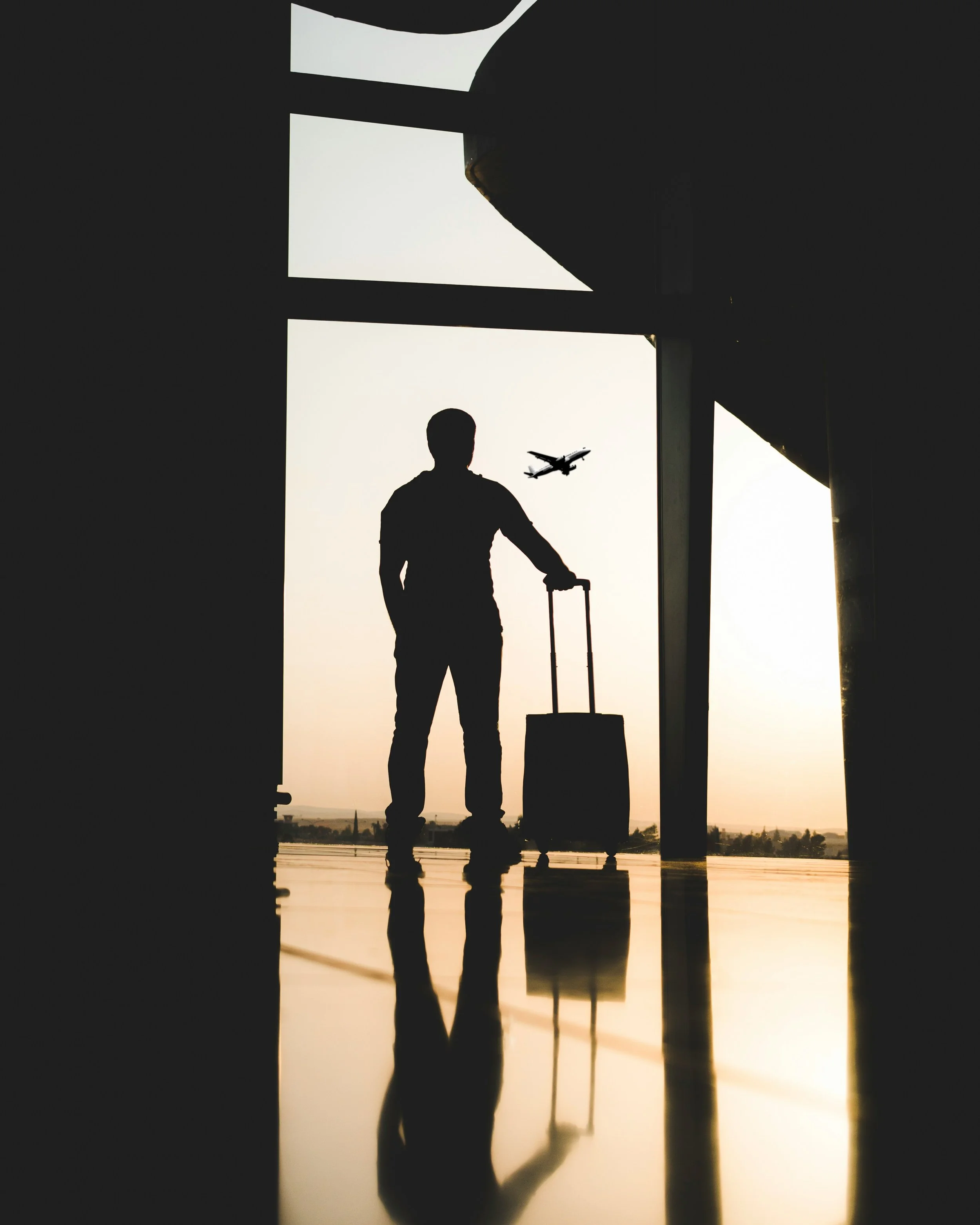 Silhouette de una persona con equipaje de mano en un aeropuerto, con un avión volando en el cielo en el fondo durante el atardecer.