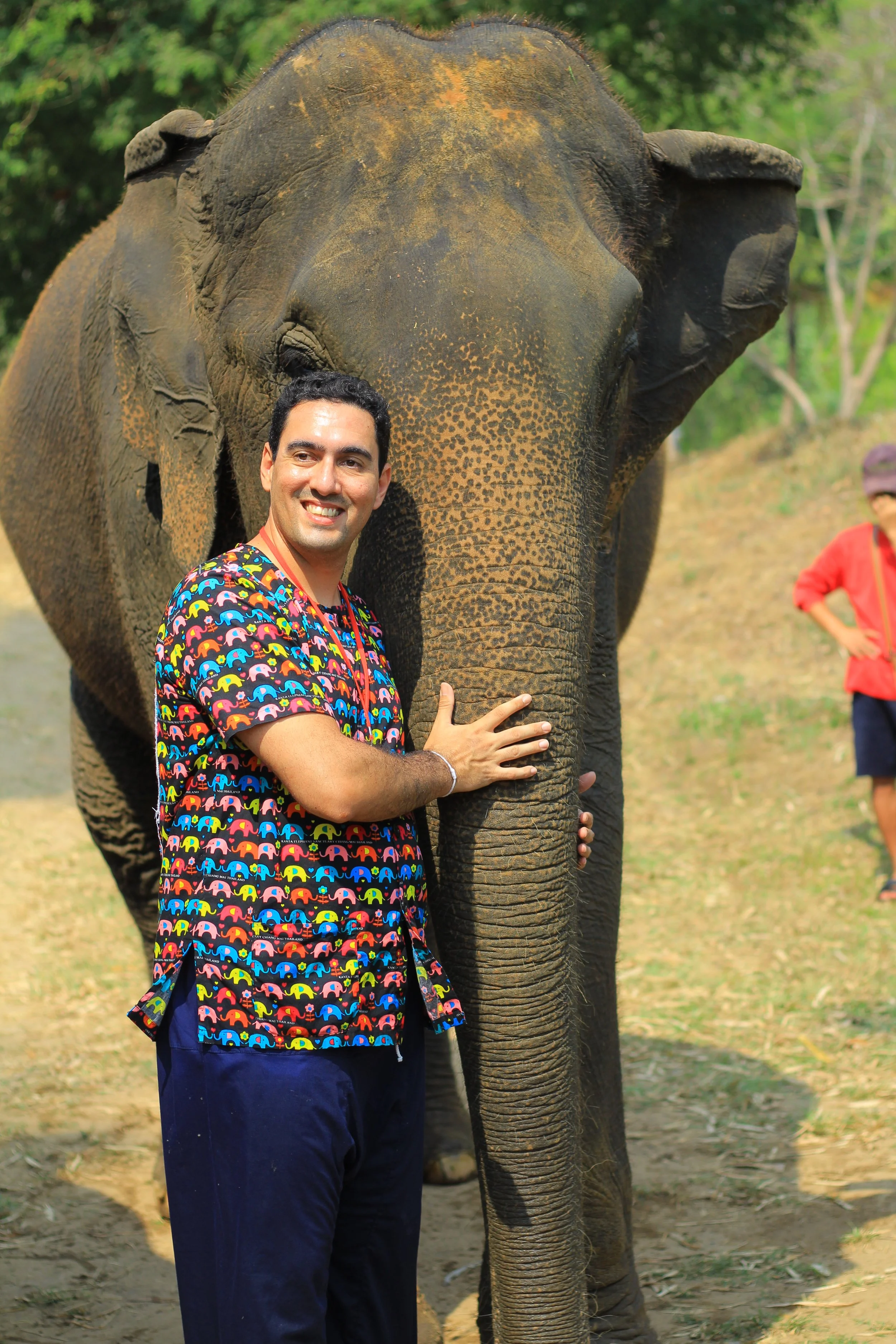 Hombre sonriendo con una camiseta con estampado de elefantes multicolores, tocando a una elefante africana en un entorno natural, con otras personas al fondo.