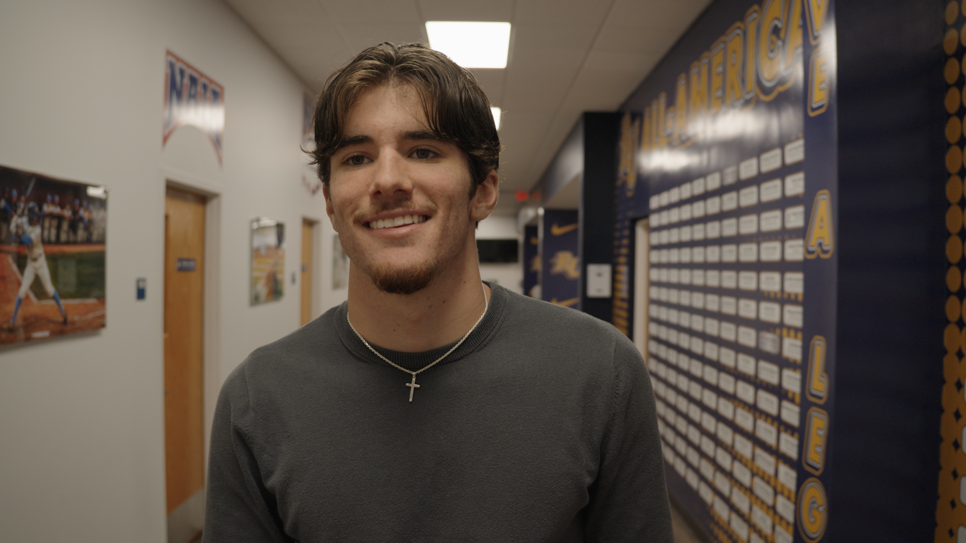Young man with brown hair, wearing a gray shirt and a silver cross necklace, standing in a hallway with sporting posters and a blue wall with a sign for the University of Georgia.