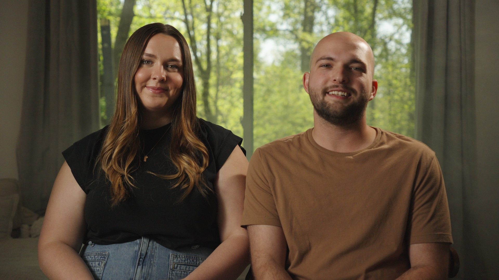 A young woman with long brown hair wearing a black top and jeans sitting beside a young man with a shaved head and a beard wearing a tan t-shirt, sitting together in front of a large window with green trees visible outside.