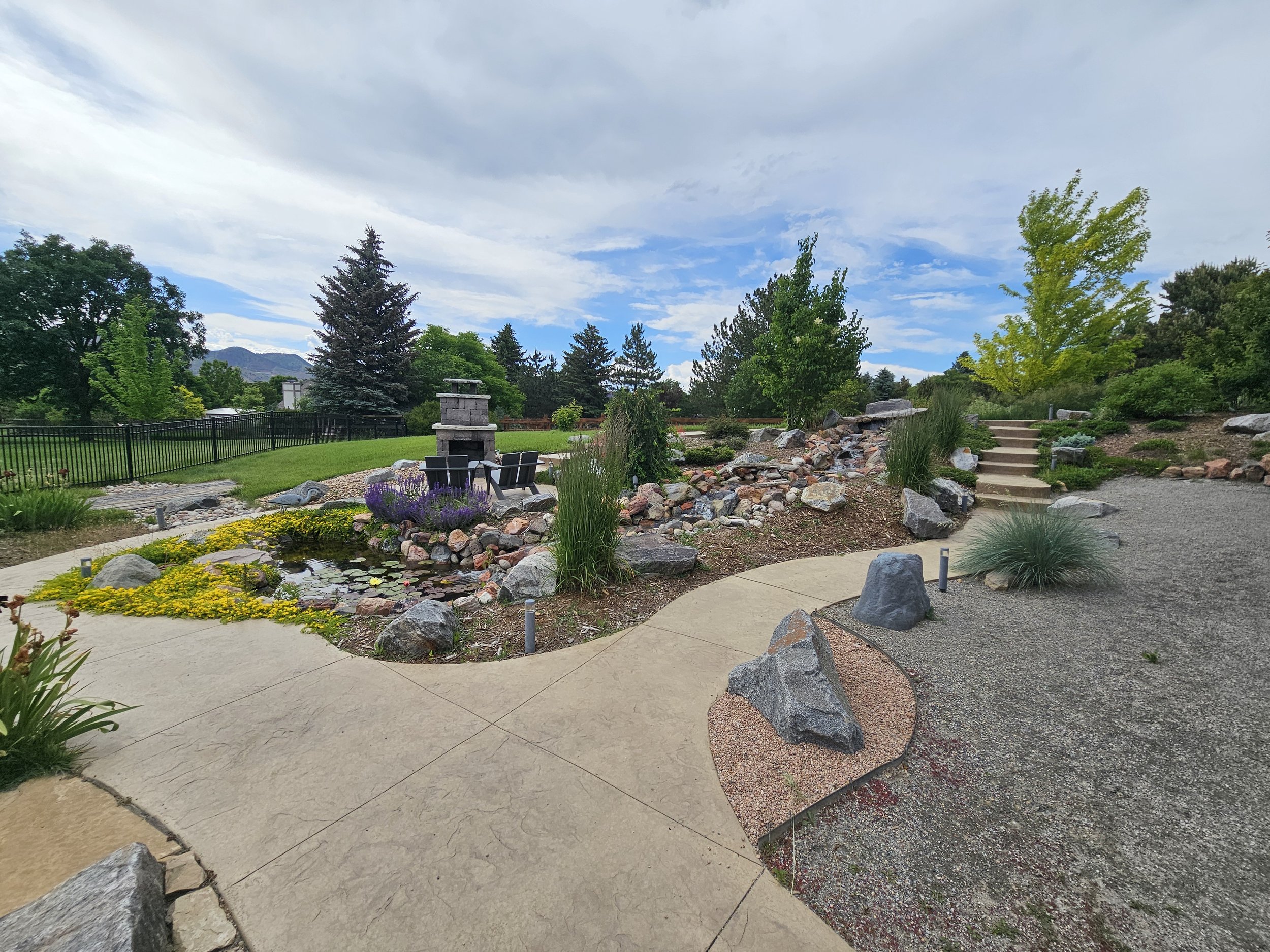 A landscaped backyard with a small pond surrounded by rocks and yellow flowers, a table with chairs next to a water feature with lavender plants, a gravel pathway, stone steps, and various green trees and bushes under a partly cloudy sky.