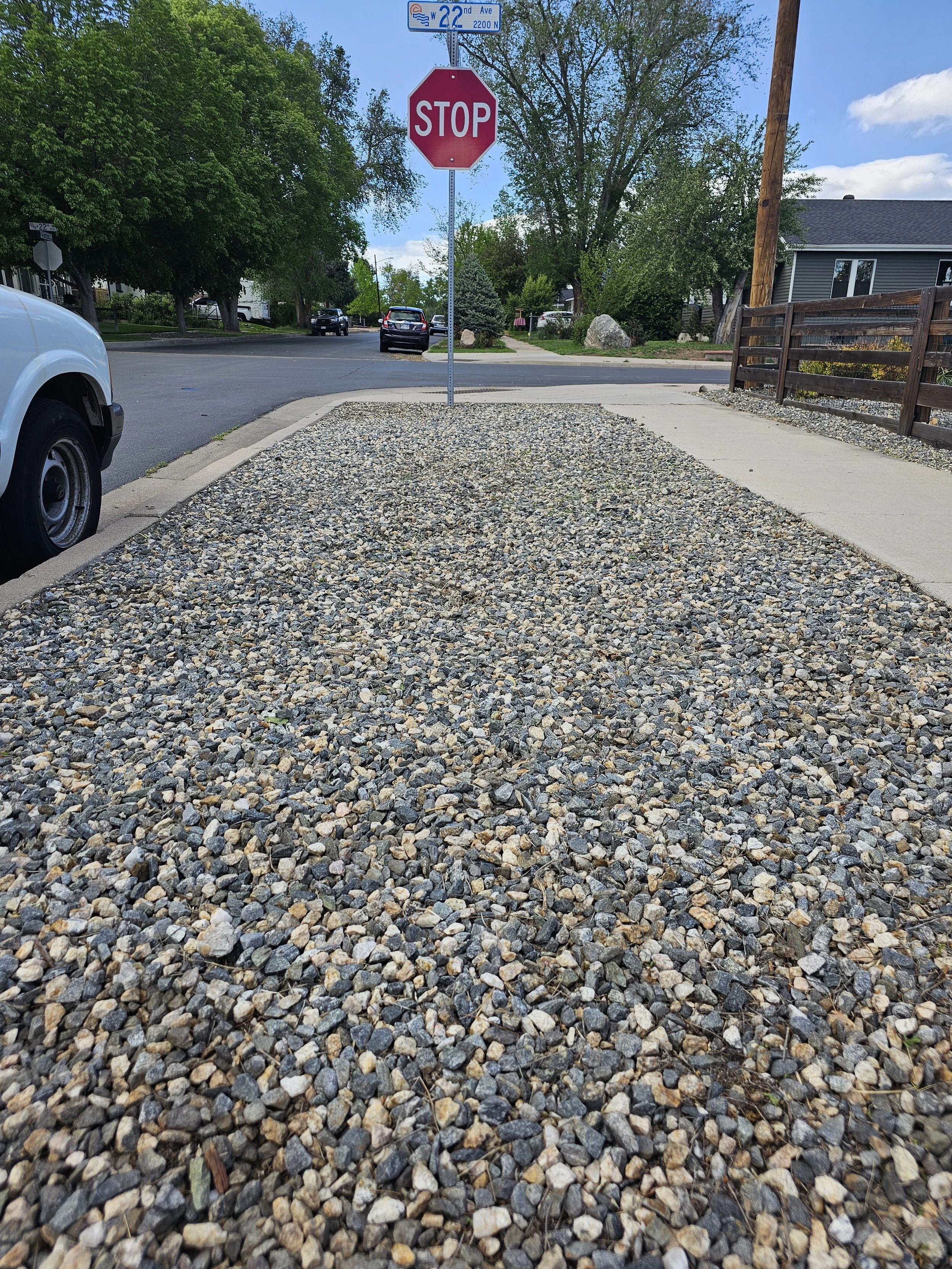 A stop sign and a street sign in a residential neighborhood with parked cars and trees, viewed from a gravel pathway. This is a photo after weeding has been done.
