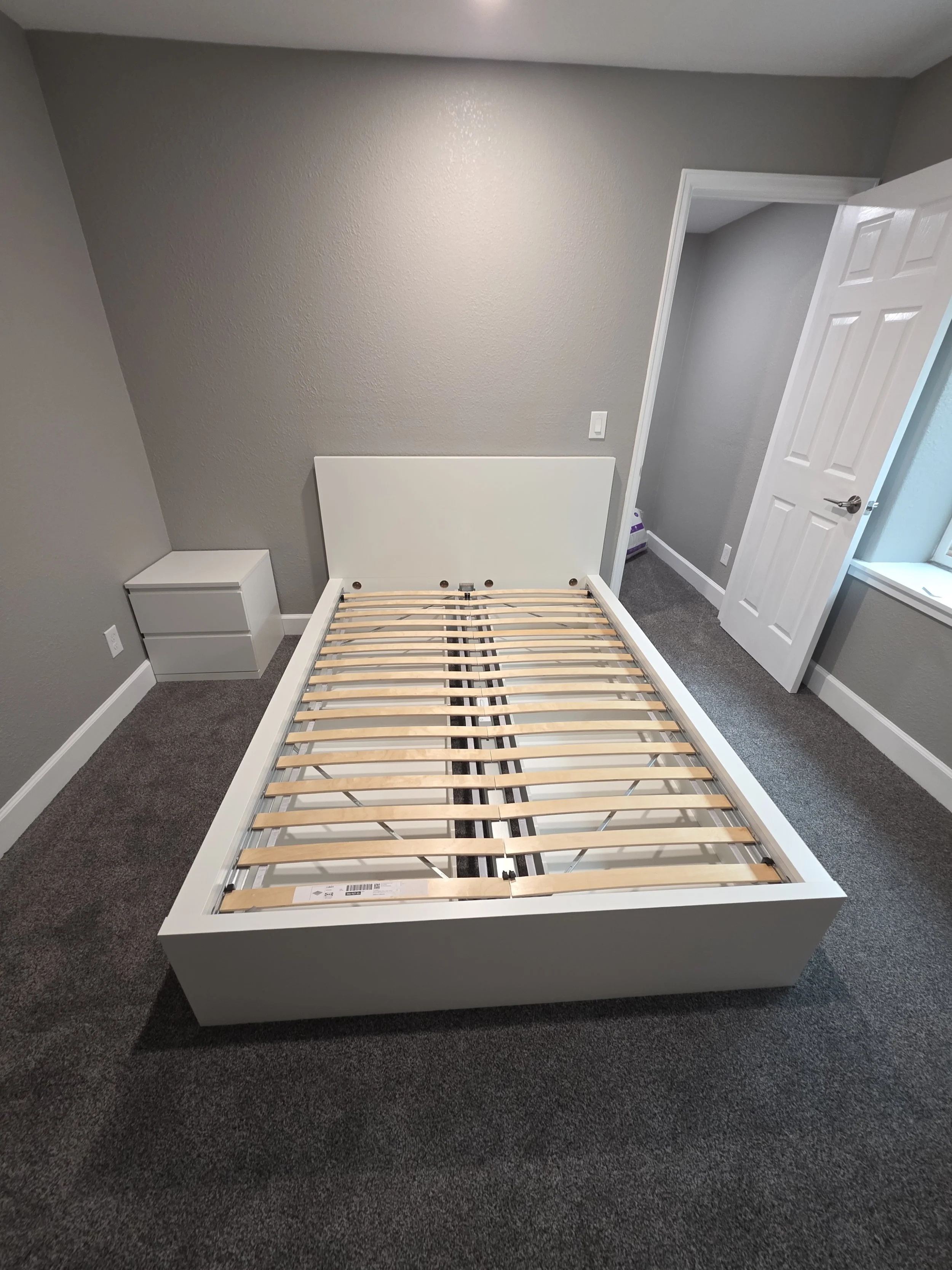 Empty white bed frame with wooden slats in a gray room, next to a small white nightstand, with an open door leading to a hallway.