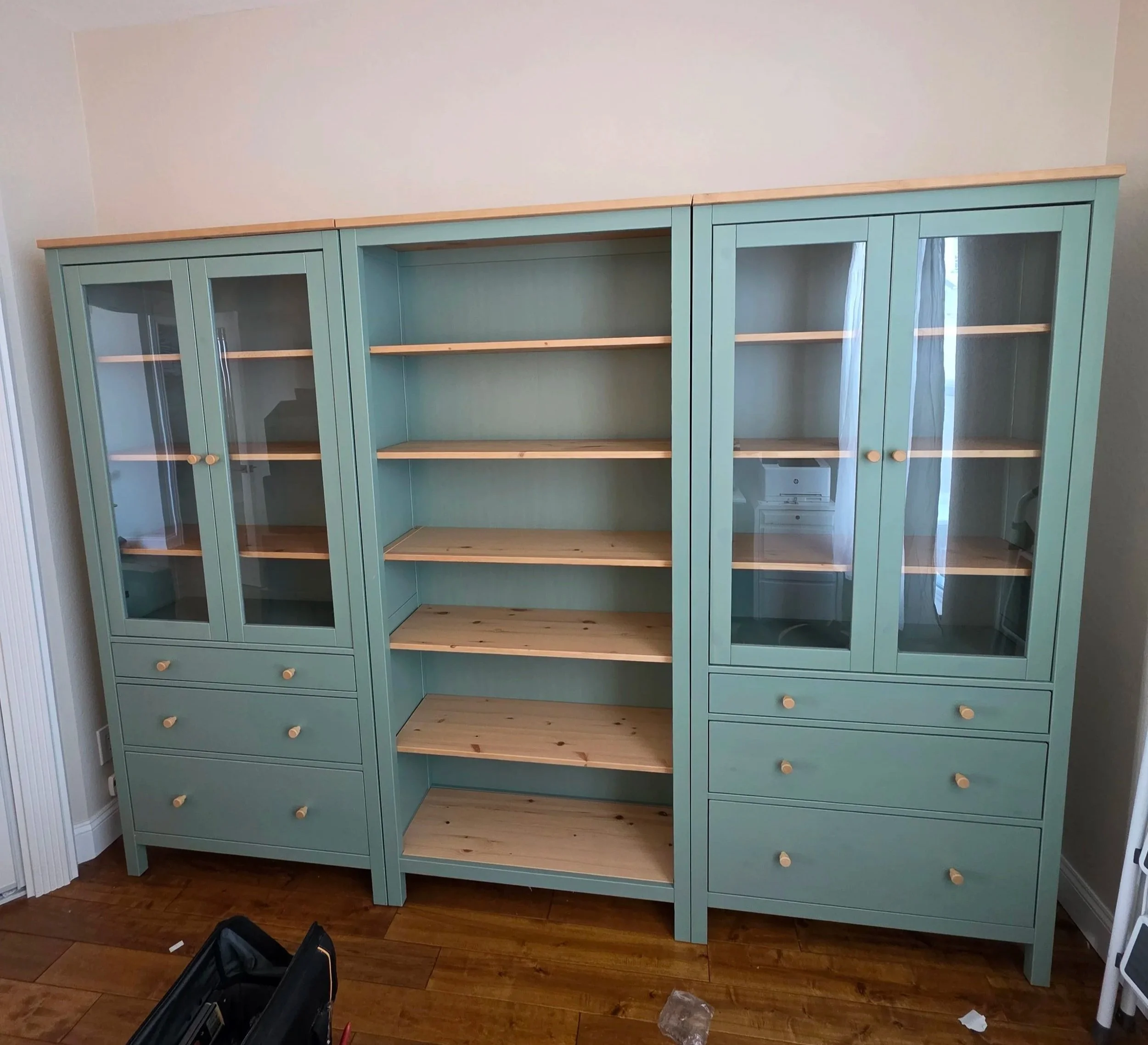 A teal wooden cabinet with glass doors and drawers is shown in an unfinished room with hardwood flooring and a plain beige wall. The cabinet has three sections, each with glass-paneled doors, and several drawers at the bottom. There are items on the floor including a black toolbox and some plastic packaging.