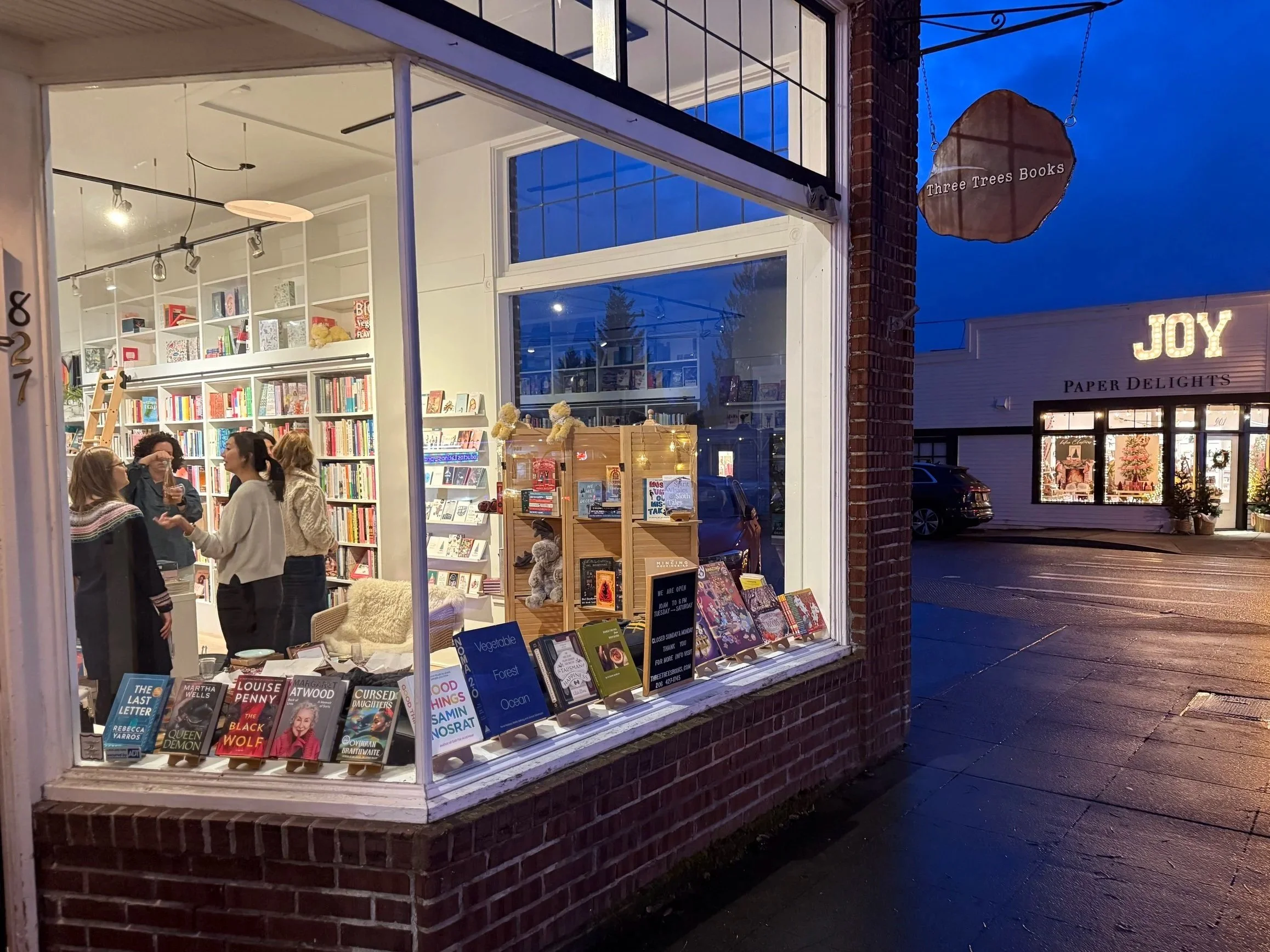 Night view of a bookstore window display with books and plush toys inside, and a street scene outside with signs for three trees books and Paper Delights, illuminated letters spelling 'JOY', and parked cars.