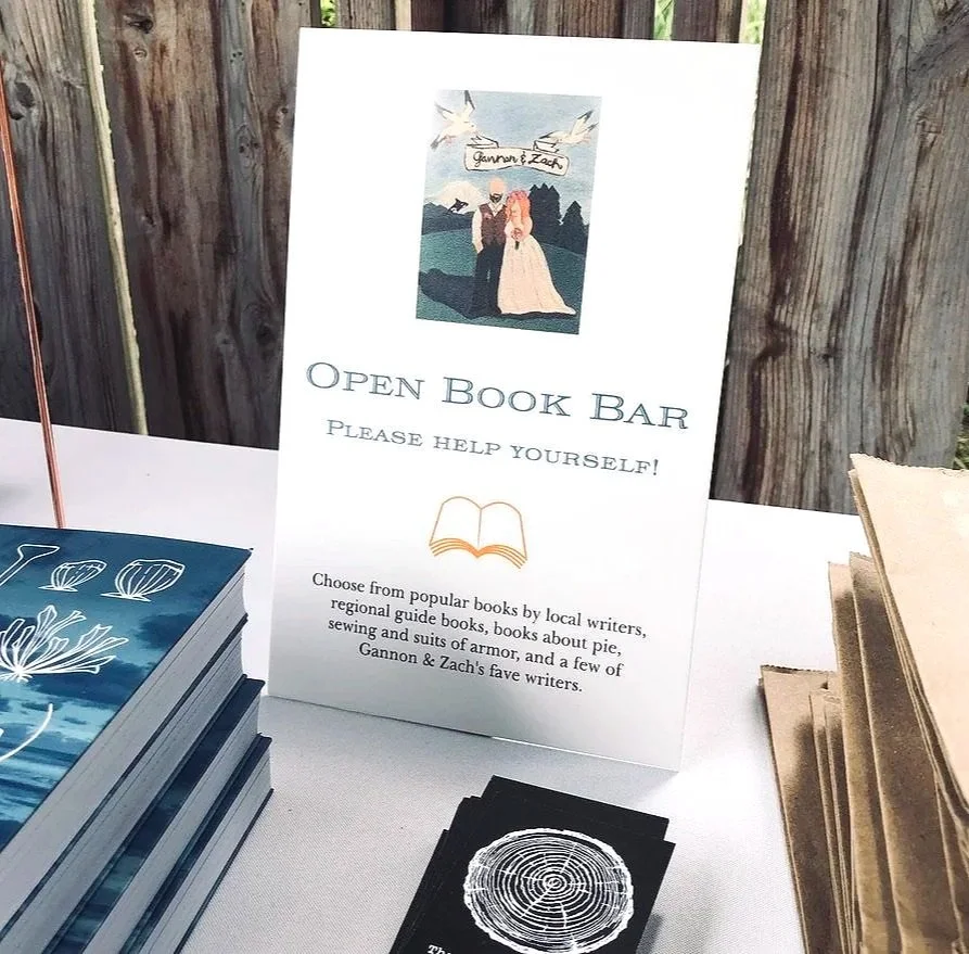 A sign for an open book bar at a table with stacks of books and a black card with a circular design.