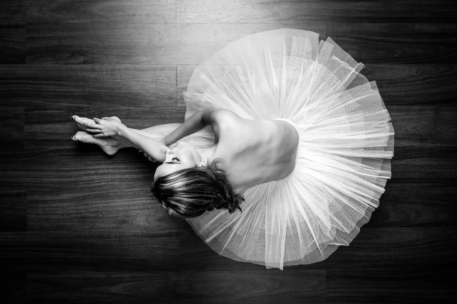 A woman in a ballet tutu lying on her side on a wooden floor, with her hands clasped and her eyes closed.