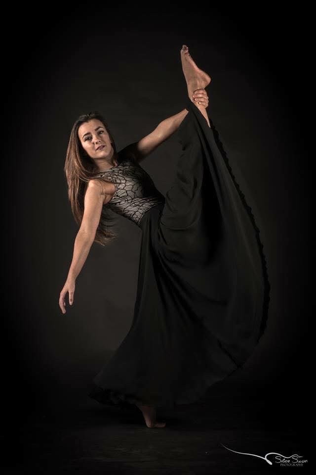 A woman in a black dress with a lace top, posing against a dark background, holding her raised leg with her hand.