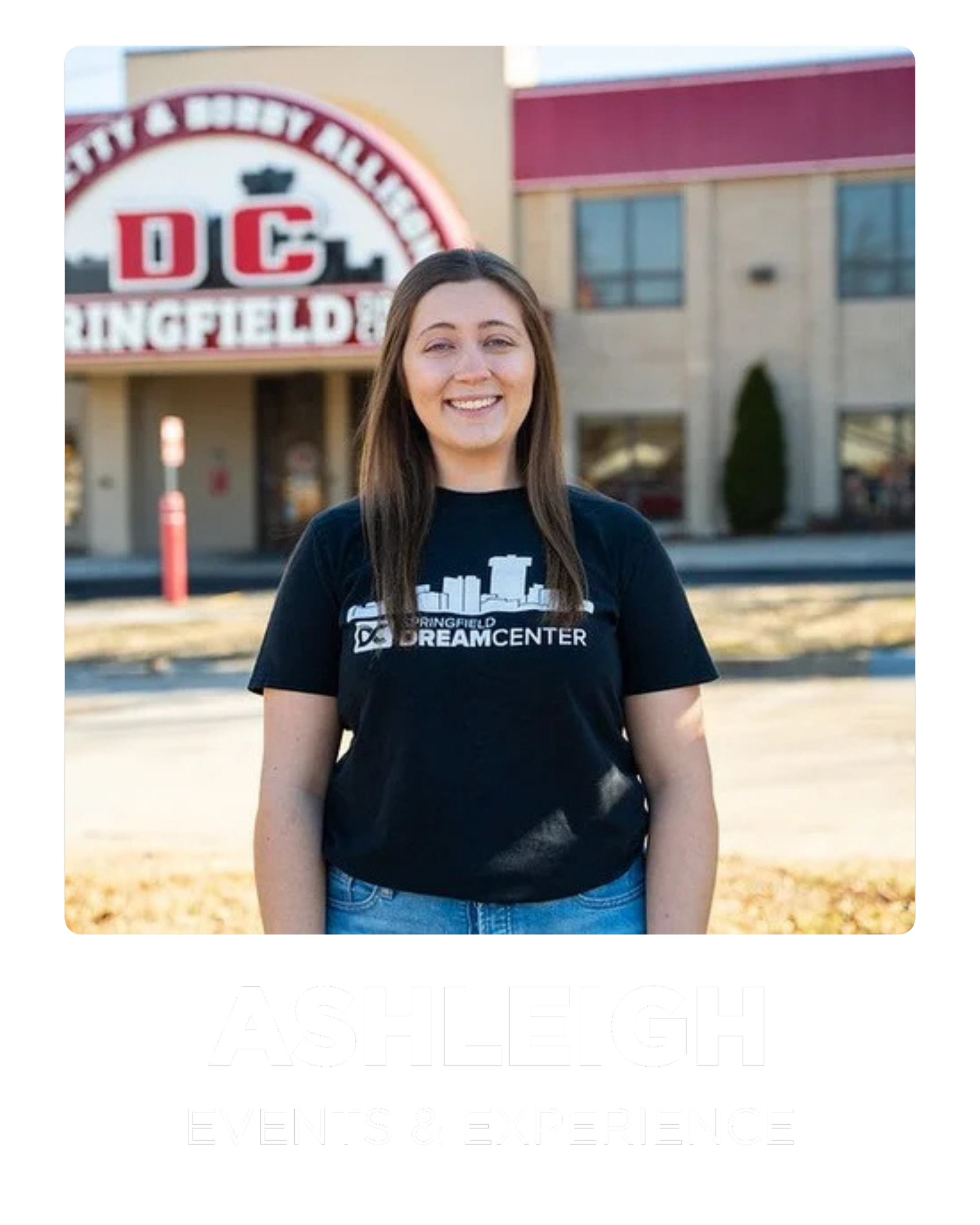A young woman standing outdoors in front of a building with a red sign that reads 'DC Springfield &'. She is smiling and wearing a black t-shirt with a city skyline graphic and the words 'Springfield Dream Center'.