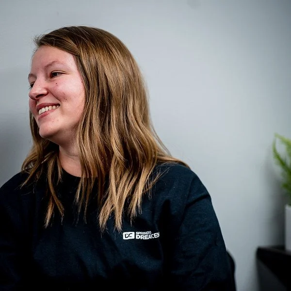Young woman with light brown hair smiling, wearing a black shirt, against a plain light gray background.
