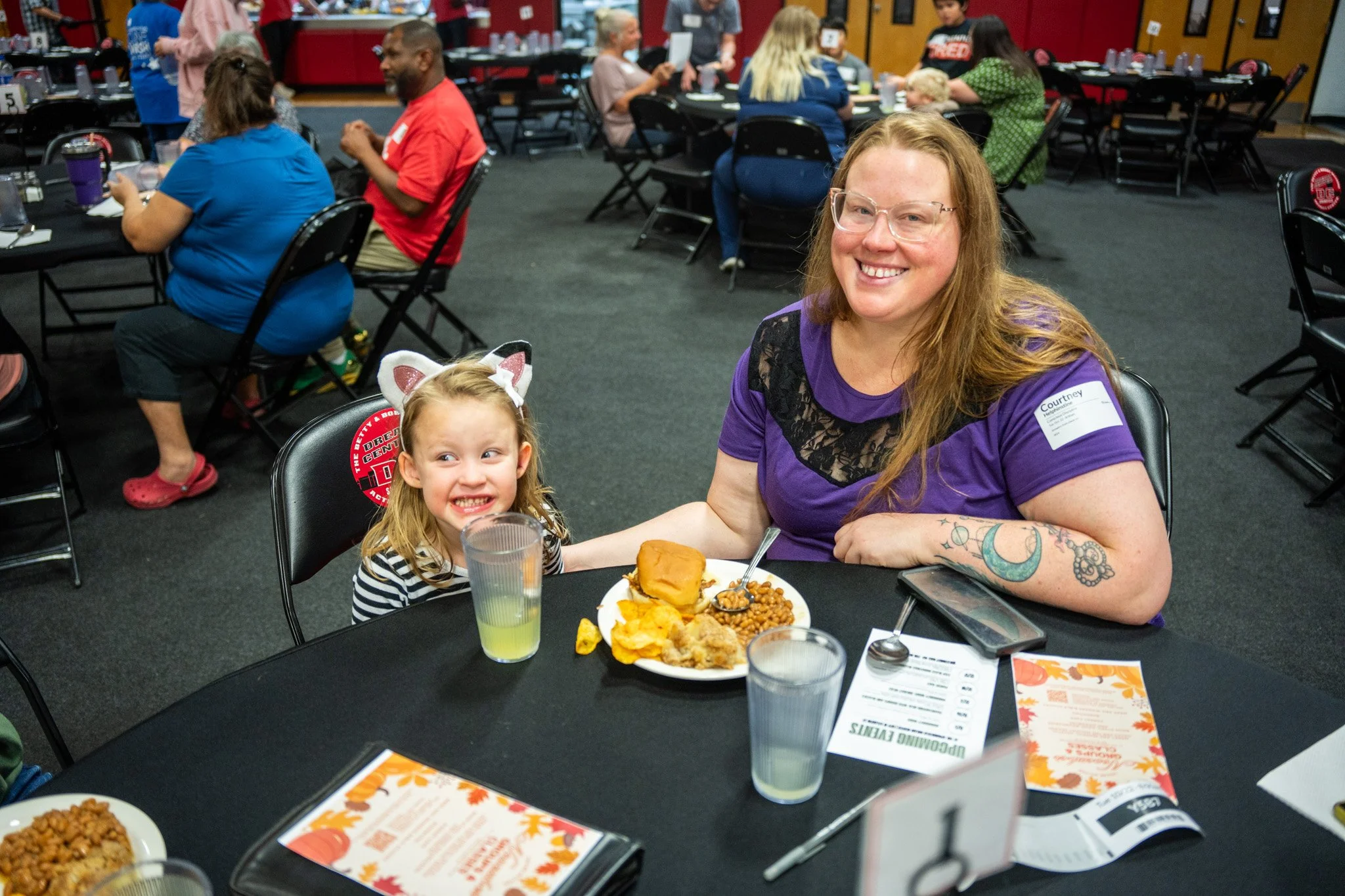A woman and a young girl sitting at a table with food, smiling at the camera in a decorated banquet hall with other guests in the background.