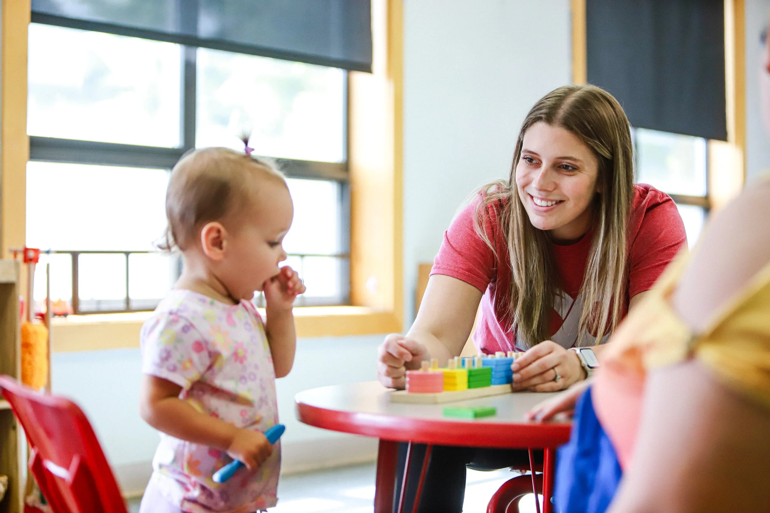 A young girl with a ponytail and pink floral shirt stands near a desk in a classroom, with her hand near her mouth. A smiling woman with long hair, wearing a red shirt, interacts with her over a small colorful game or puzzle on the desk. The background shows windows with black blinds and a yellow window frame, indicating a bright classroom setting.