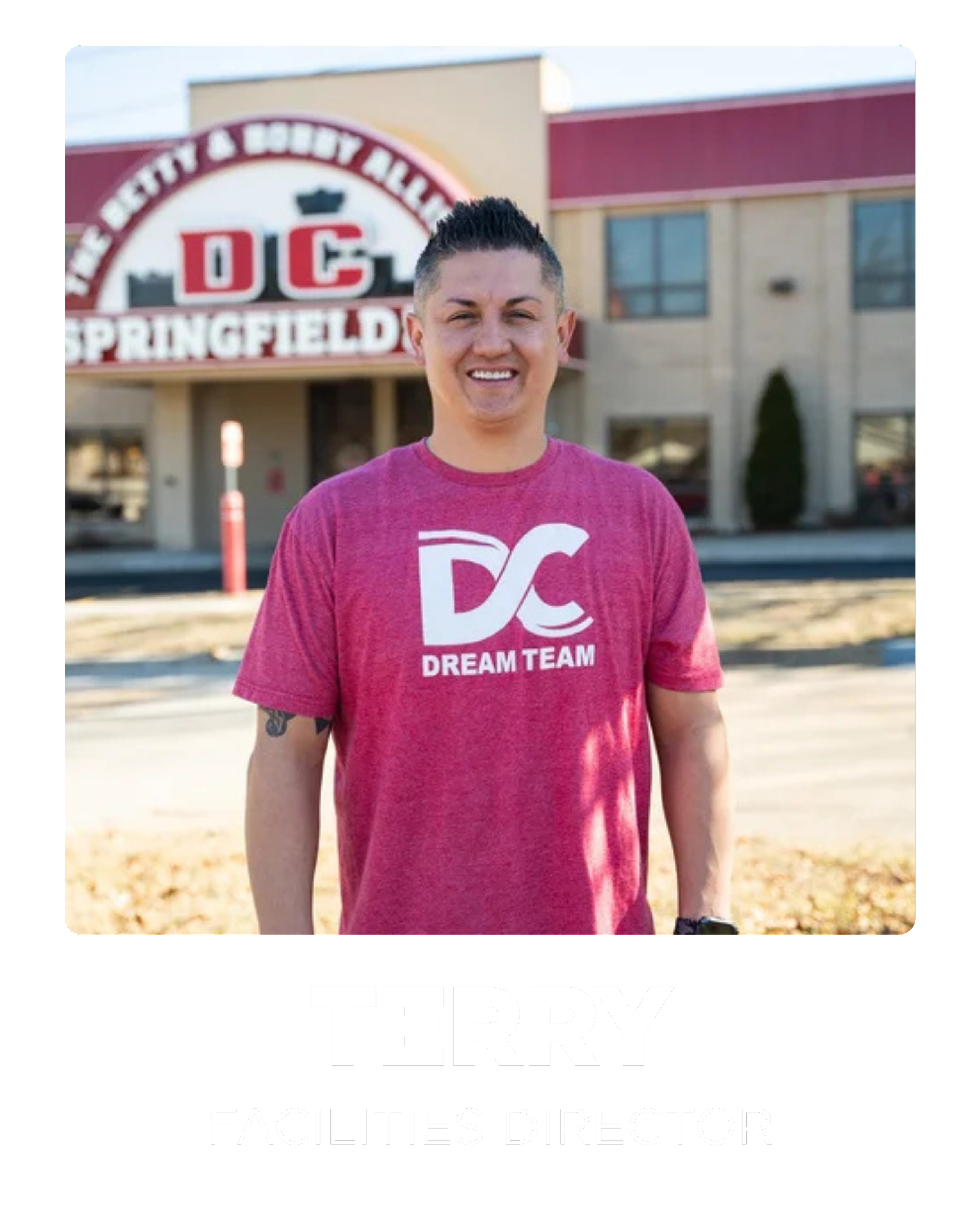 A man standing outside a building with a sign that reads 'DC Springfield.' He is wearing a pink T-shirt that says 'DC Dream Team' and is smiling at the camera.