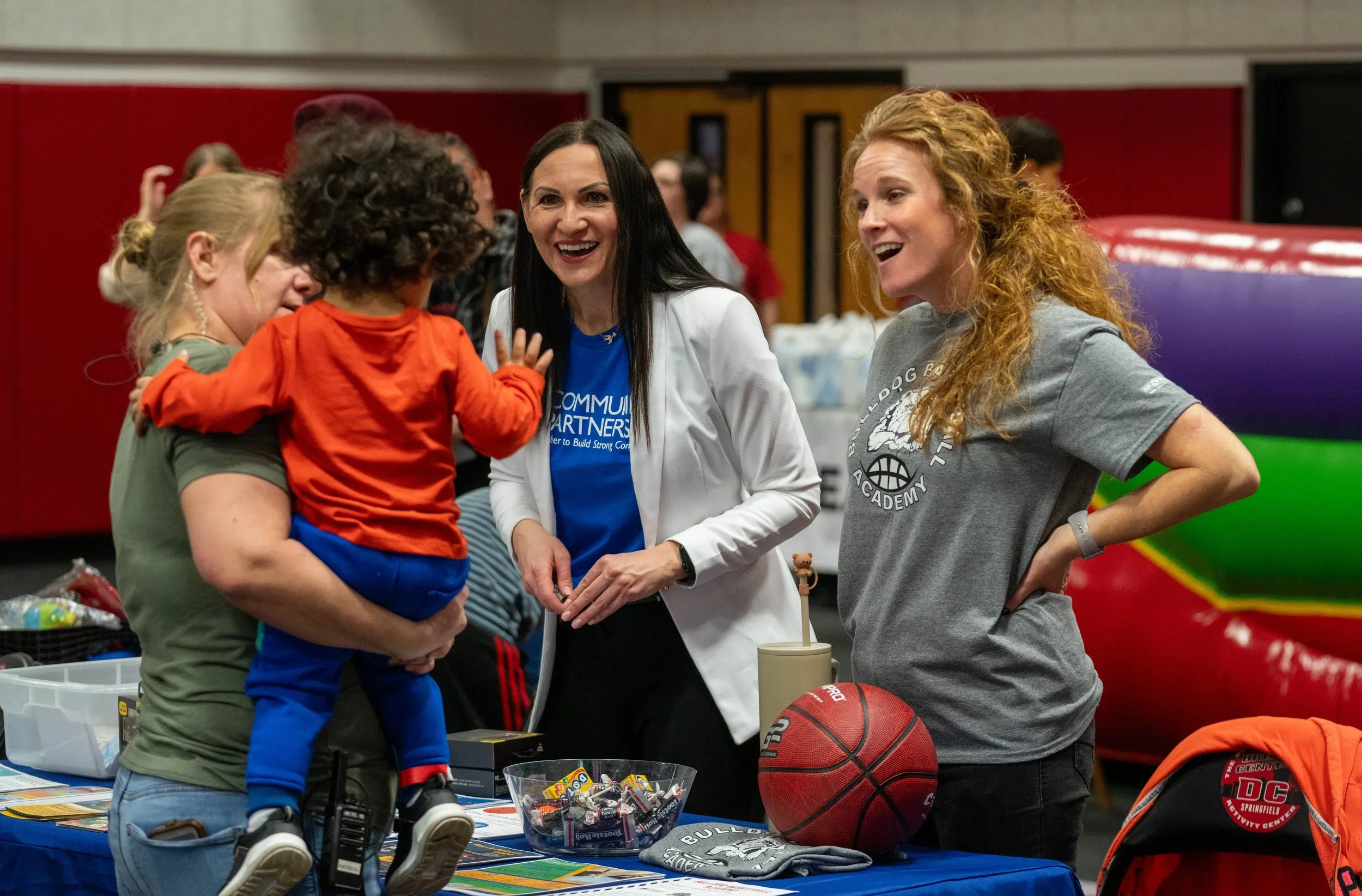 Two women and a child at a community event. The woman on the left holds a child in an orange shirt, while the woman in the white jacket and blue shirt is talking to a woman in a gray T-shirt. The table has a basketball, a bowl of candy, and other items.