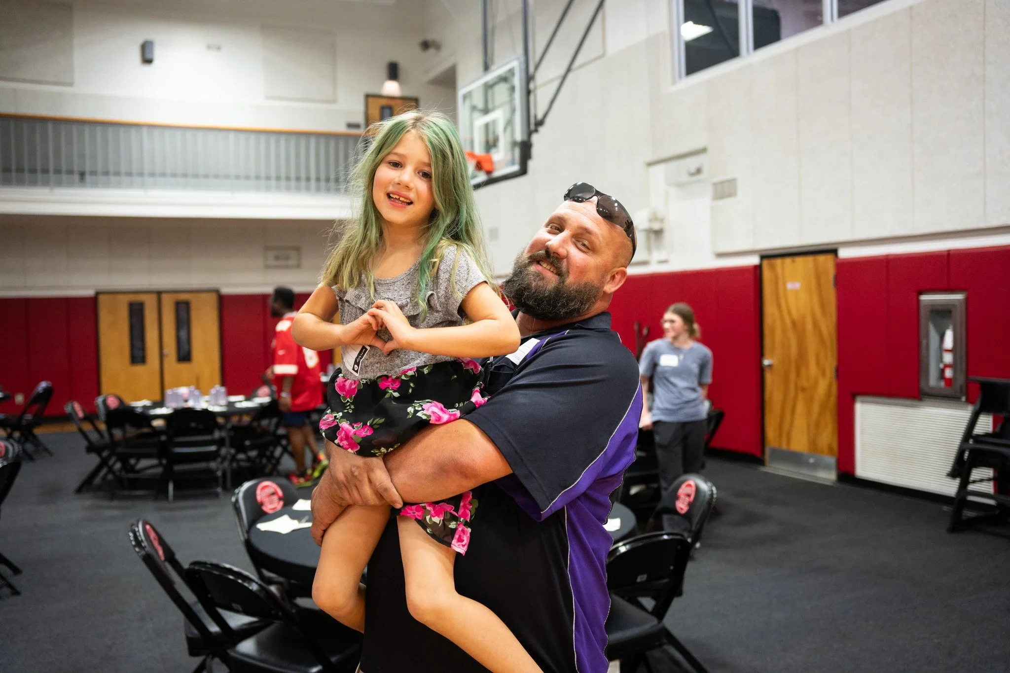 A man with a beard and sunglasses holding a young girl with green hair at a social gathering in a gymnasium, with tables and people in the background.