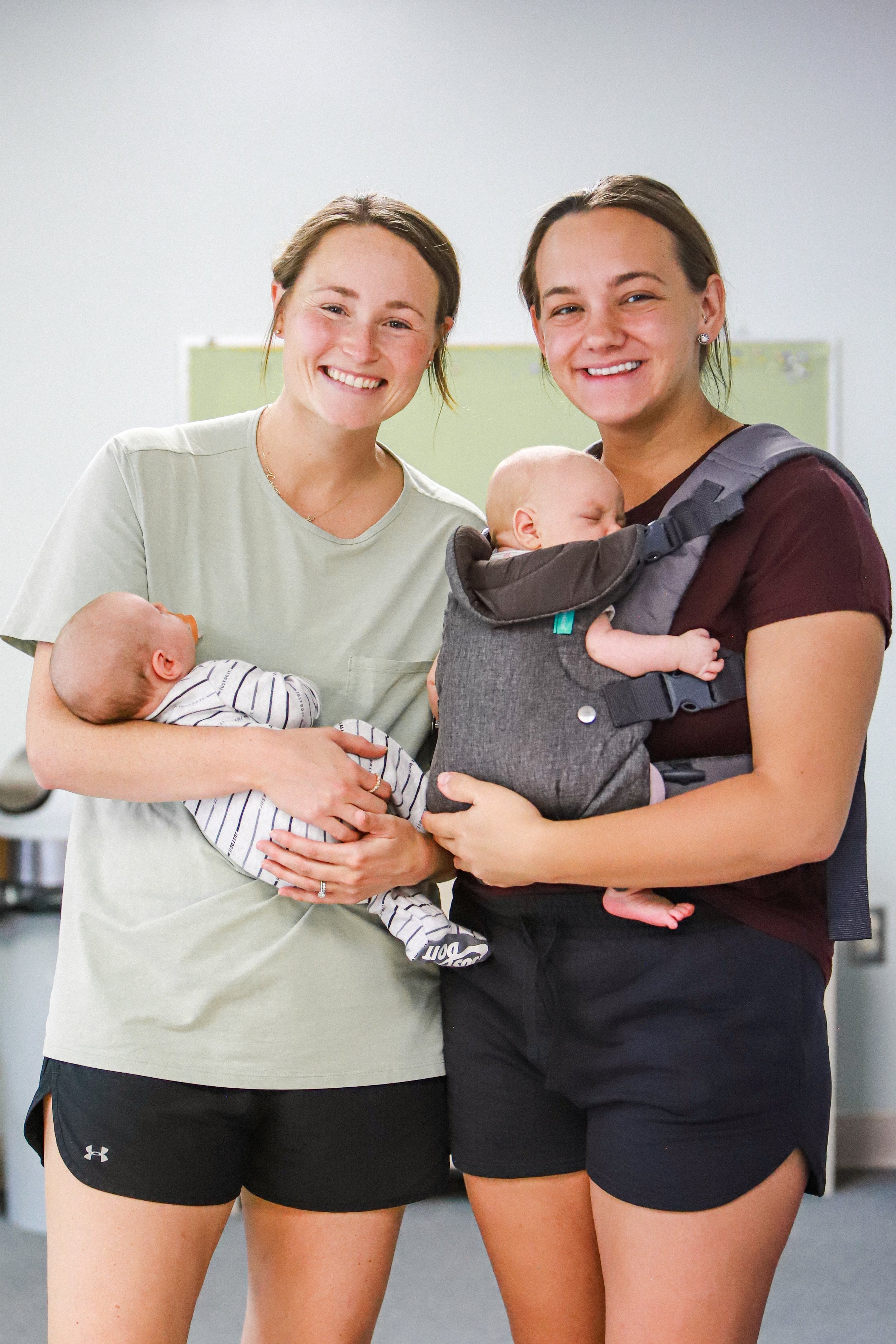 Two smiling women holding sleeping babies. One woman is wearing a light green T-shirt and black shorts, and the other is wearing a dark red T-shirt and black shorts. The babies are dressed in striped onesies.