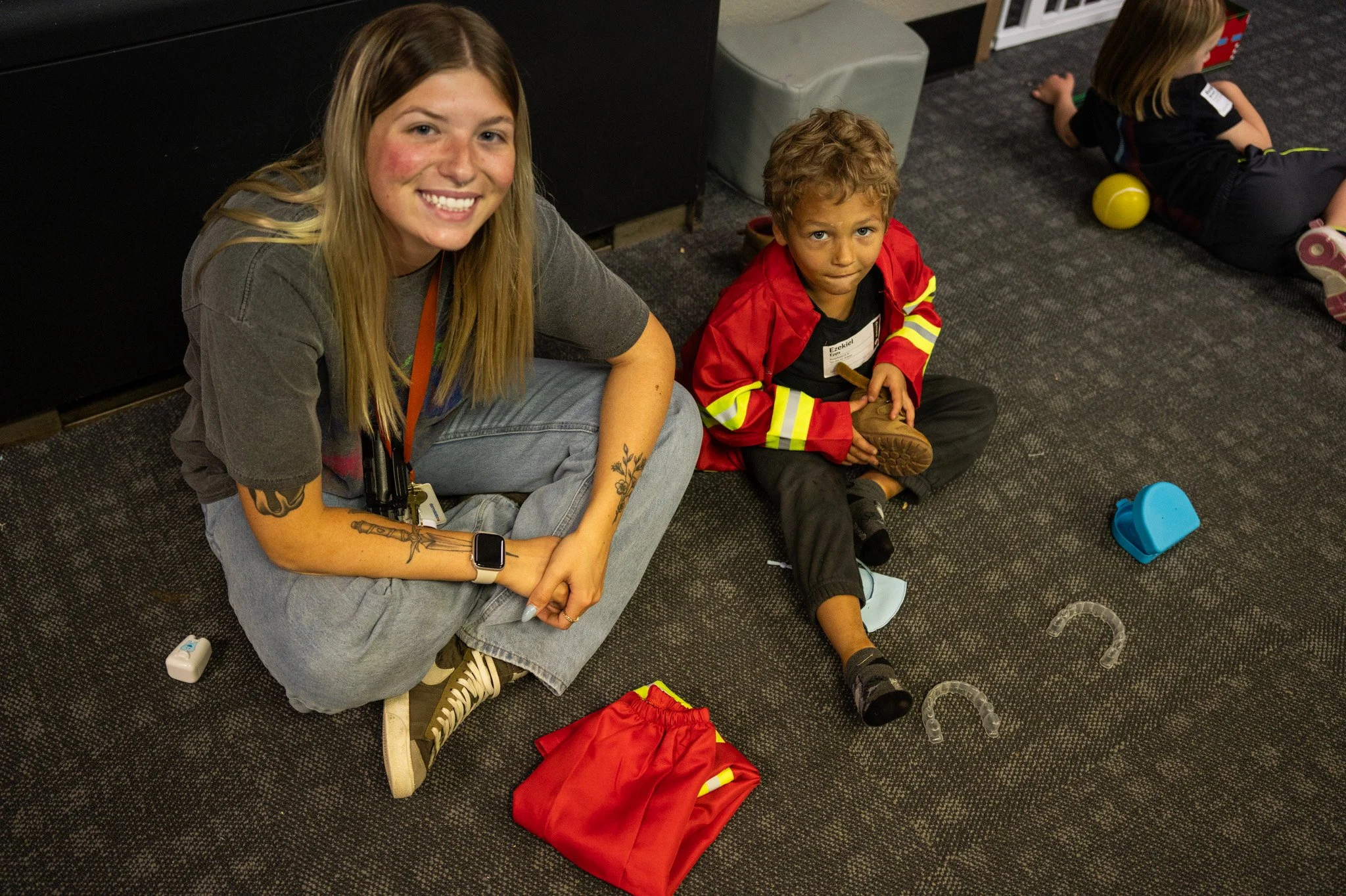 A young woman with tattoos on her arm sitting next to a boy in a firefighter costume on a black carpeted floor, smiling at the camera. There are toys and a red costume on the ground and another child in the background.