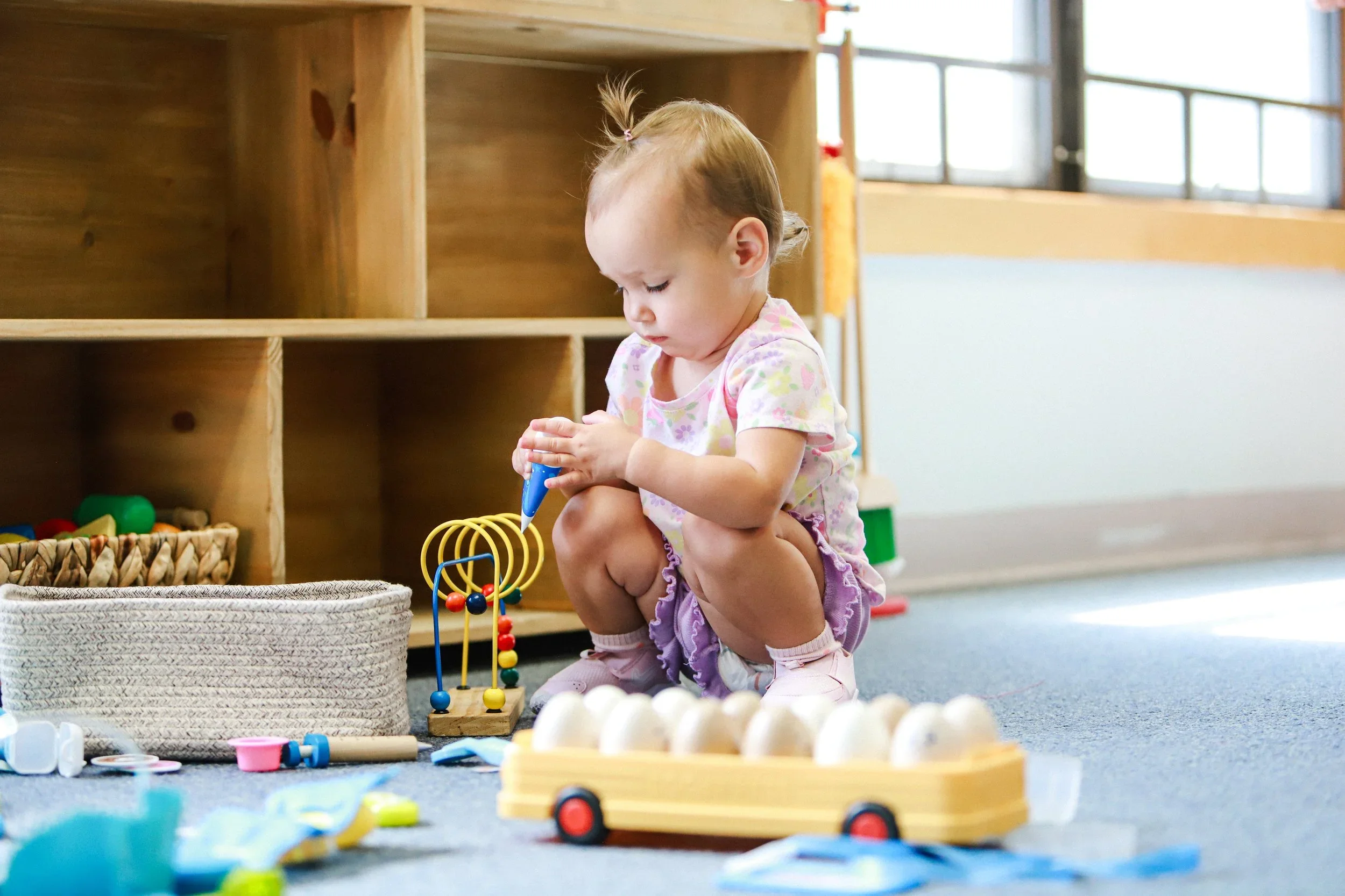 Young girl playing with colorful wooden and plastic toys on the carpeted floor in a playroom with wooden shelves and large windows.