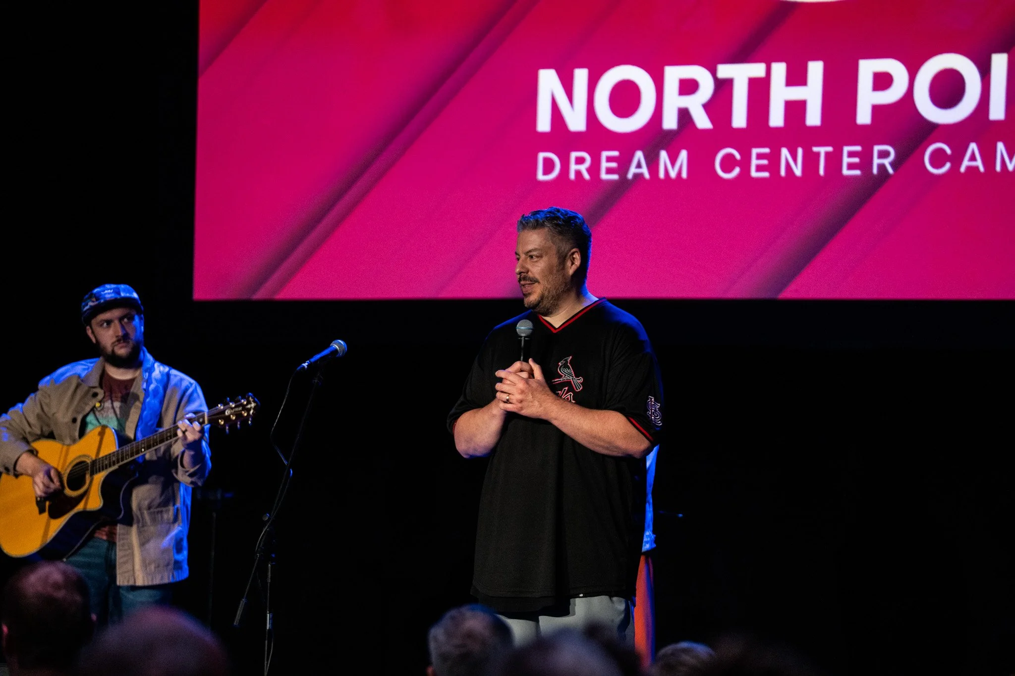 A man holding a microphone speaking on stage with a musician playing guitar beside him during the North Point Dream Center campaign event.