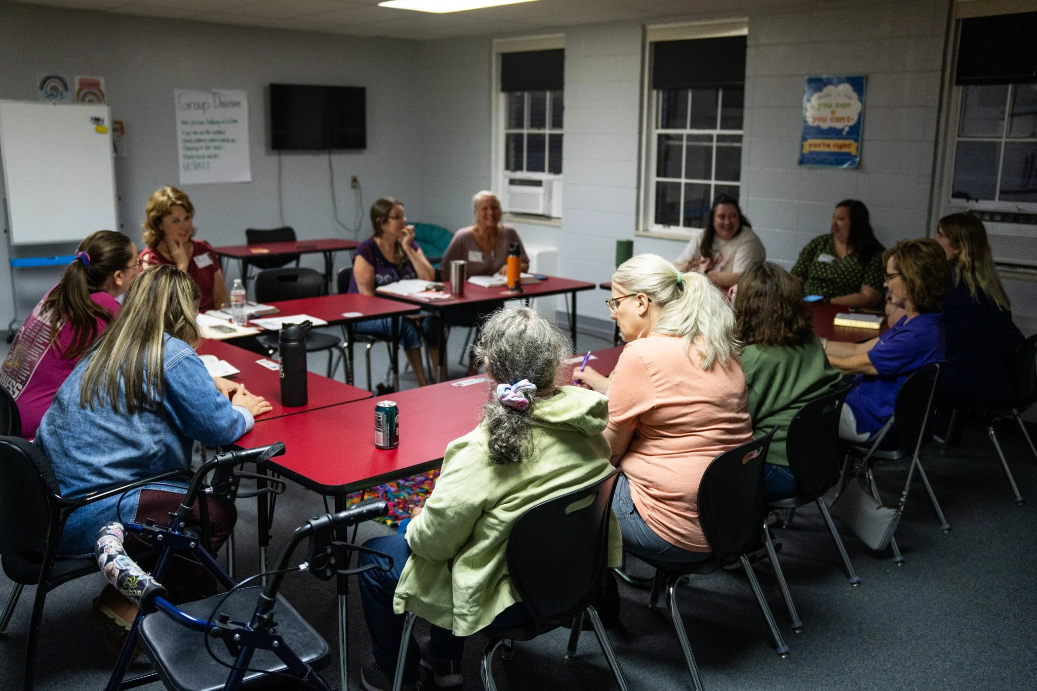 A group of women sitting around red conference tables in a classroom or meeting room, engaged in discussion. The room has white walls, windows, and some posters on the walls.