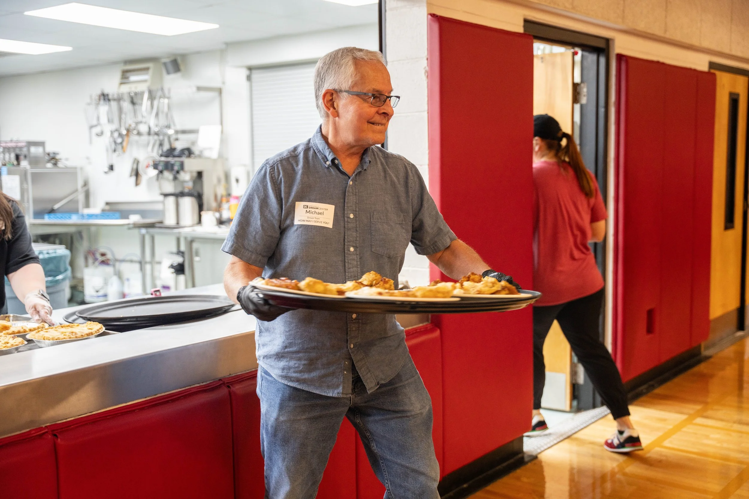 A man wearing glasses, a gray short-sleeve shirt, and gloves, holding a tray of food at an indoor event. The man has a name tag that reads 'Michael' and is standing near a kitchen counter with another person preparing food in the background.