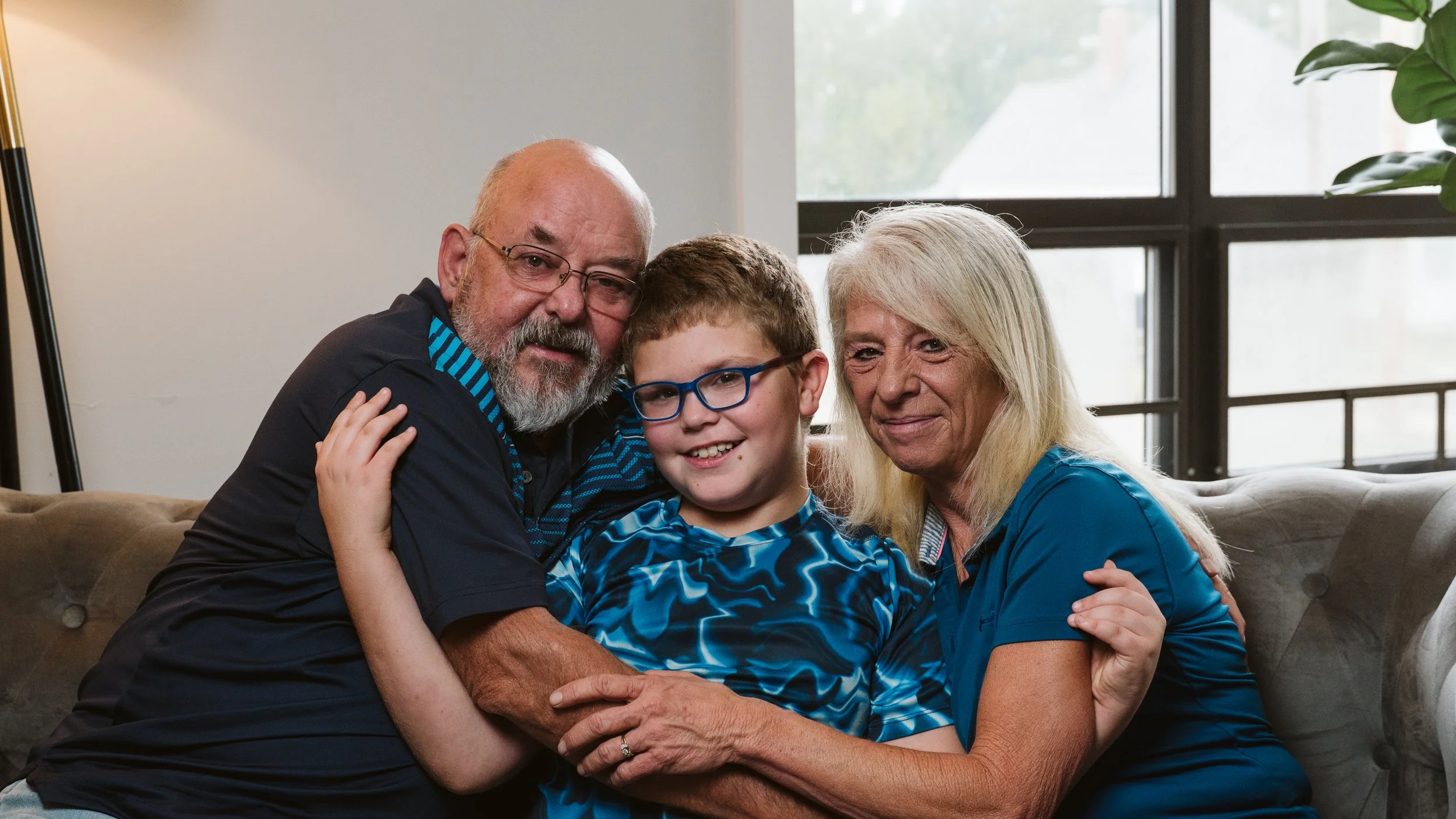 A family of three, including an elderly man and woman, and a young boy, sitting on a beige couch in a well-lit room with large windows, smiling and hugging.