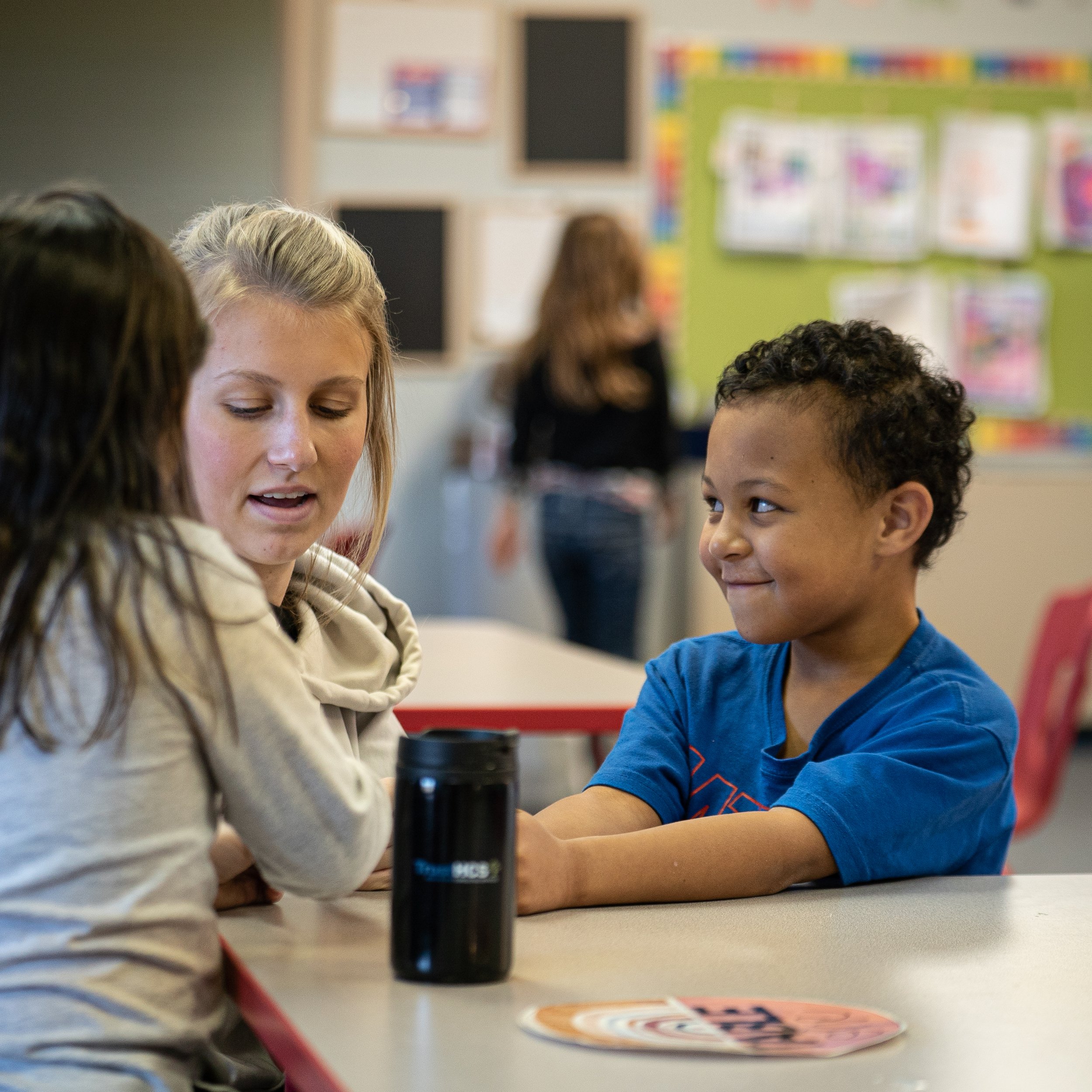 A classroom scene with a woman and two children sitting at a table, engaging in conversation. The background features classroom decorations and a bulletin board.
