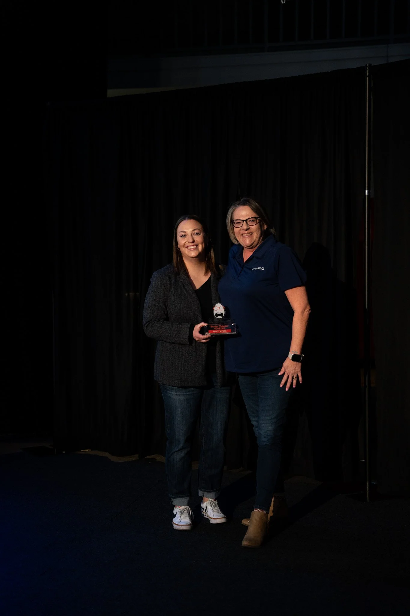 Two women standing on a stage, smiling, with one holding an award. Dark curtain backdrop.