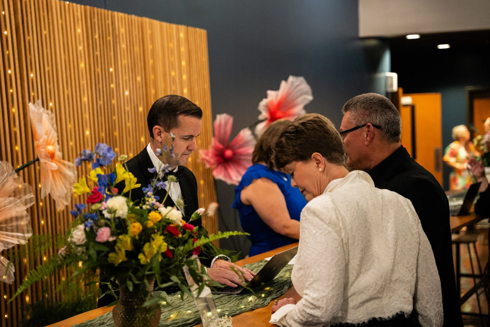 People gathered at a reception desk with a woman in white, a woman in blue, and two men in black tuxedos, one using a laptop. Decor includes a large bouquet of colorful flowers and large pink tissue paper flowers on the wall.