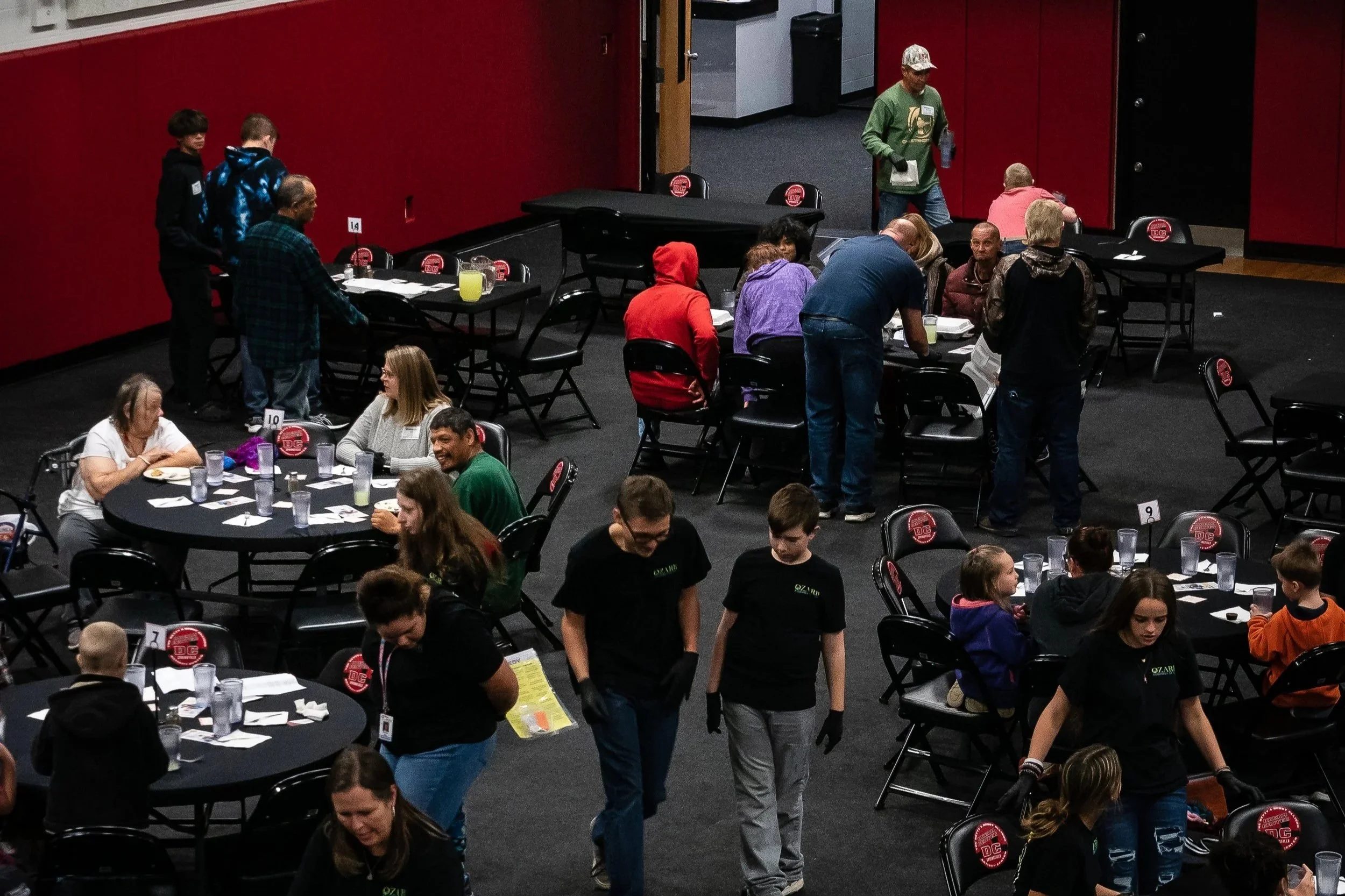 People gathered at tables inside a venue, some standing and some seated, with a red wall and black chairs, some tables are numbered, and a person in a green hoodie is walking with a water bottle in hand.
