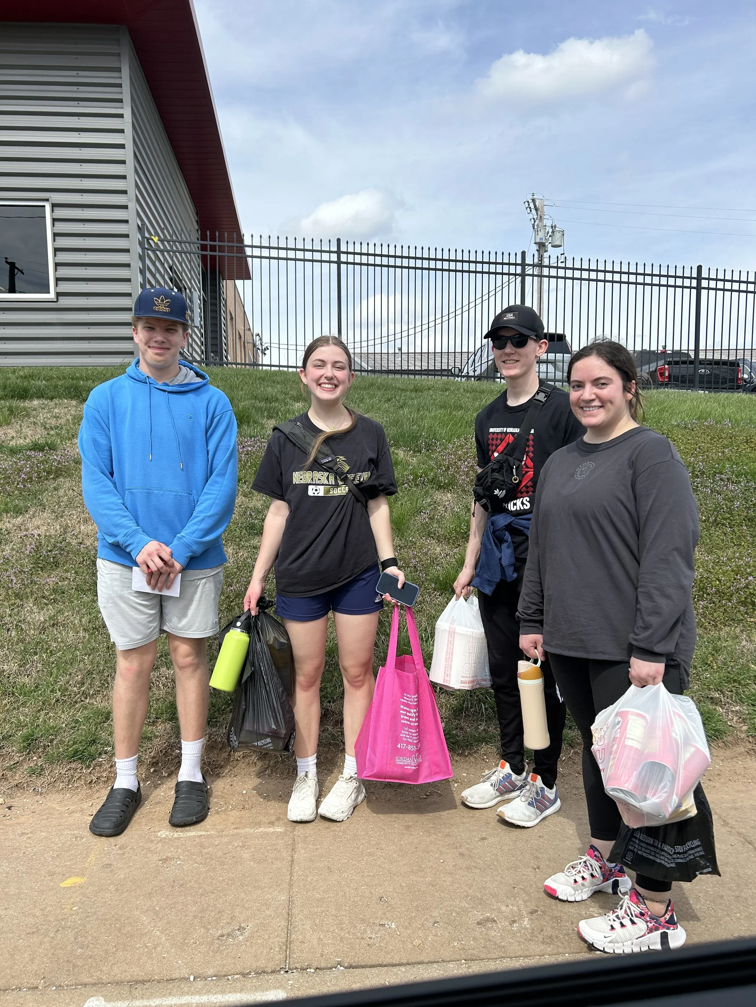 Four teenagers standing outdoors on a sidewalk, smiling and holding shopping bags and bottles.