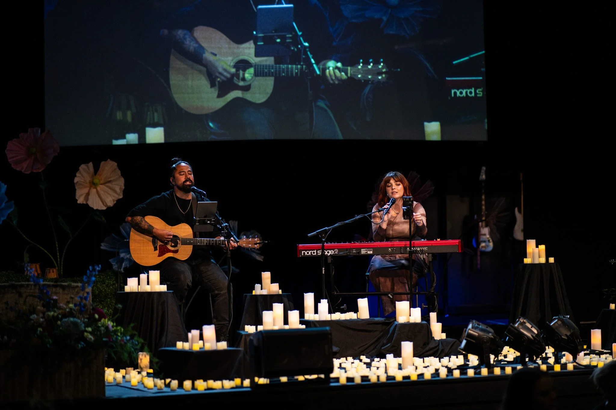 A musical performance on stage with two singers, a man with a guitar, and a woman with a keyboard, surrounded by numerous candles. A large screen above shows a close-up of the guitarist.