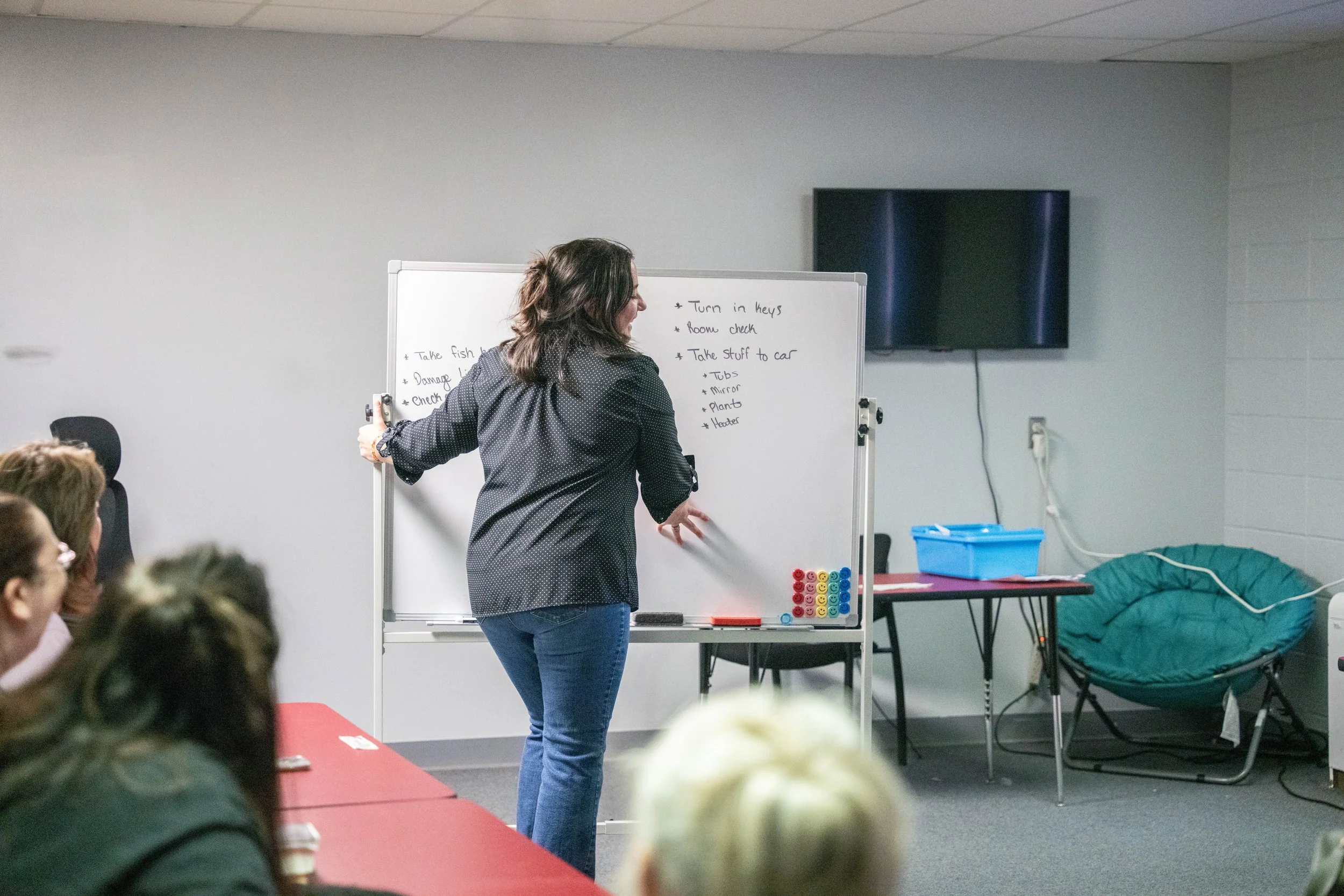 A woman writes on a whiteboard during a classroom or training session, with several students seated at a red table in front of her. A television is mounted on the wall to her right, and a blue container and a green reclining chair are visible in the background.