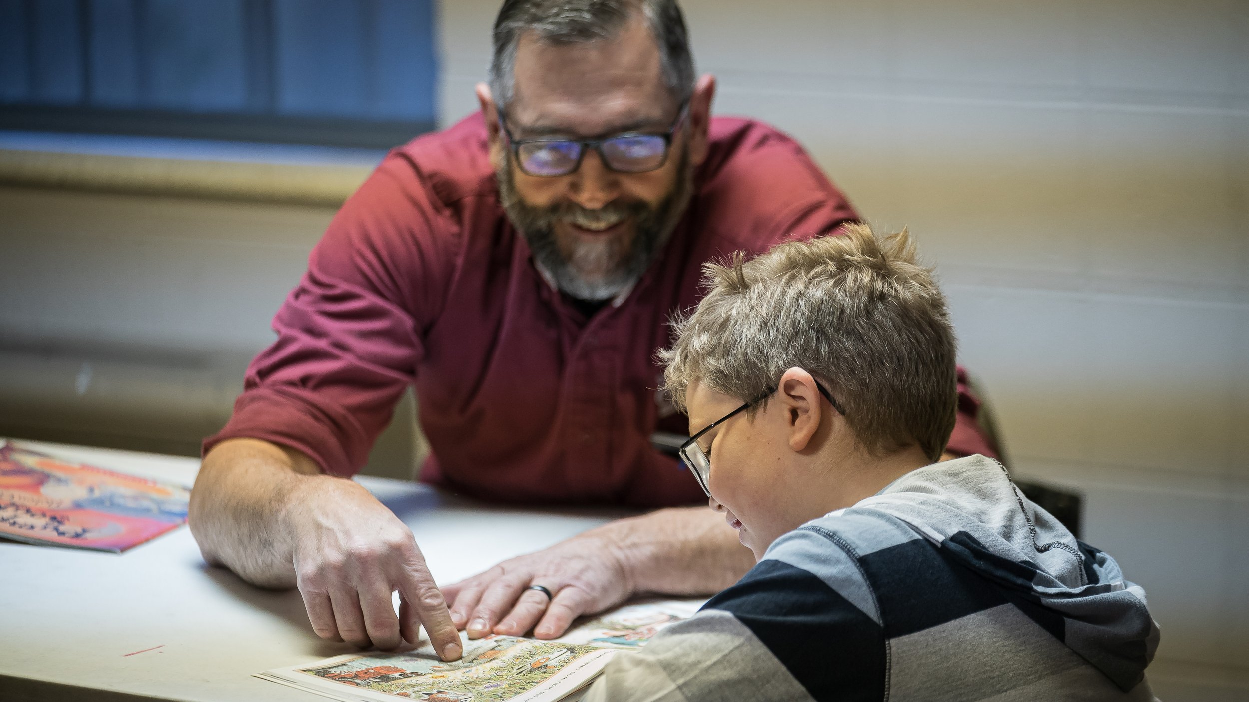 An older man with glasses and a beard, wearing a red shirt, is pointing at a comic book or illustrated story while a young boy with glasses and a striped hoodie is looking at it, sitting at a table.