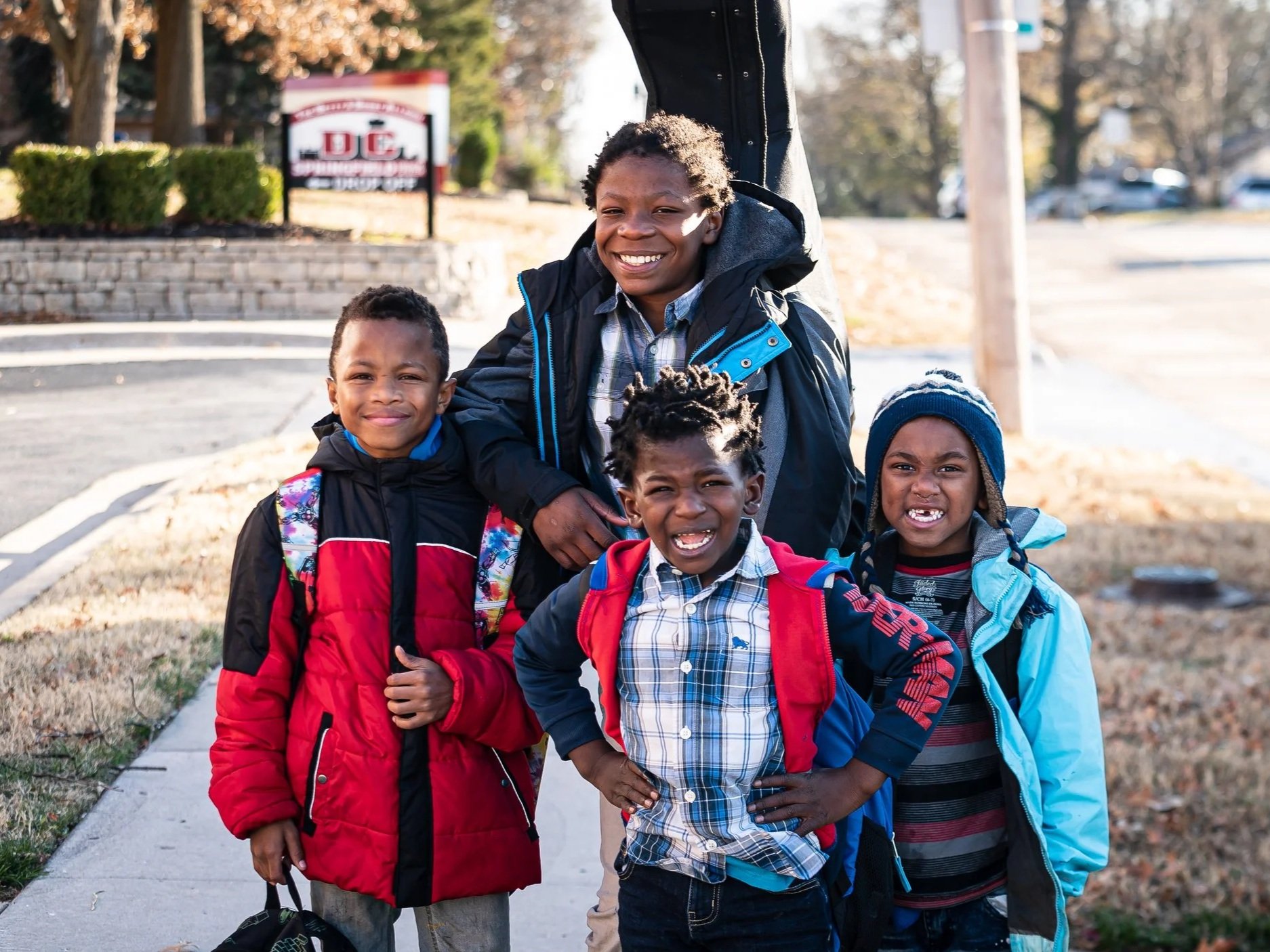 A group of five children smiling outdoors, wearing colorful jackets and backpacks, standing on a sidewalk with autumn trees and a sign in the background.