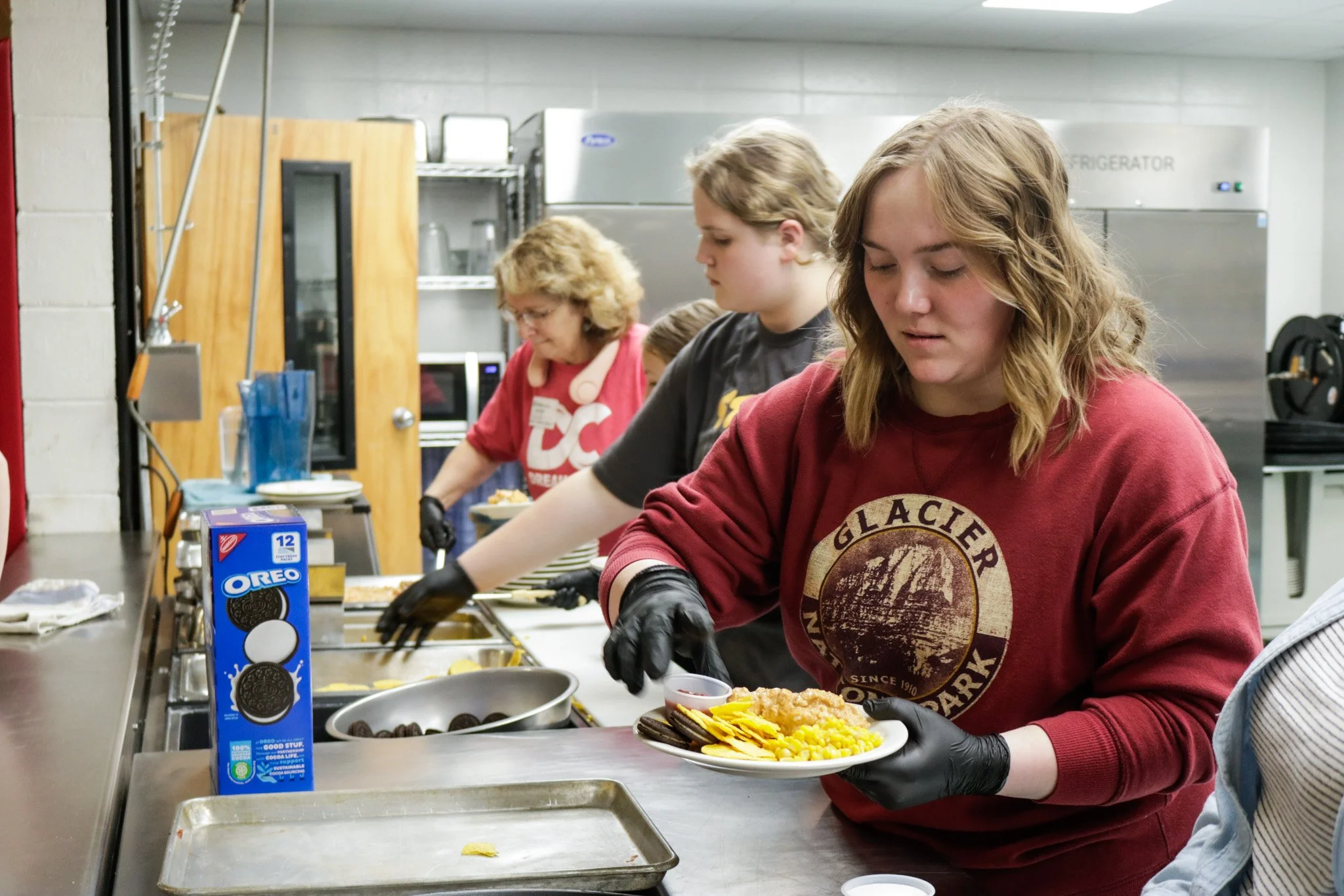 People serving and preparing food in a kitchen during a food service event.