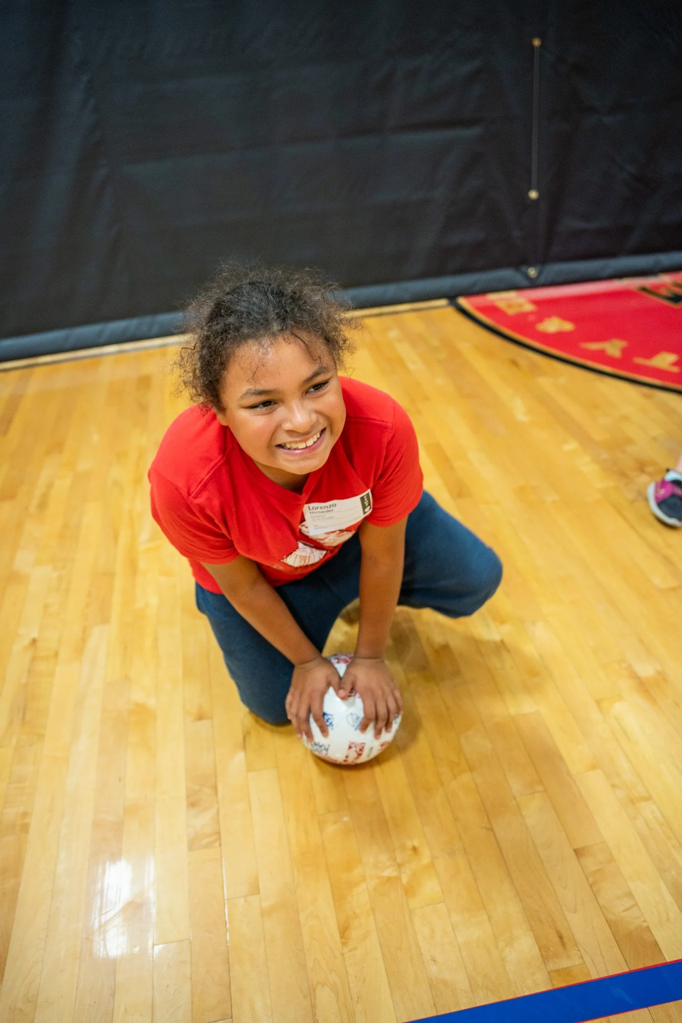 A young girl with curly hair wearing a red t-shirt and dark blue jeans, kneeling on a wooden gymnasium floor, holding a volleyball with a smile on her face.