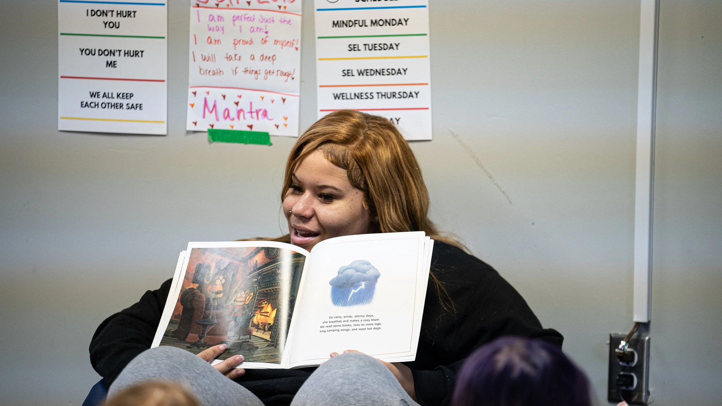A young woman with reddish hair reading a children's book about weather to children in a classroom or library setting.