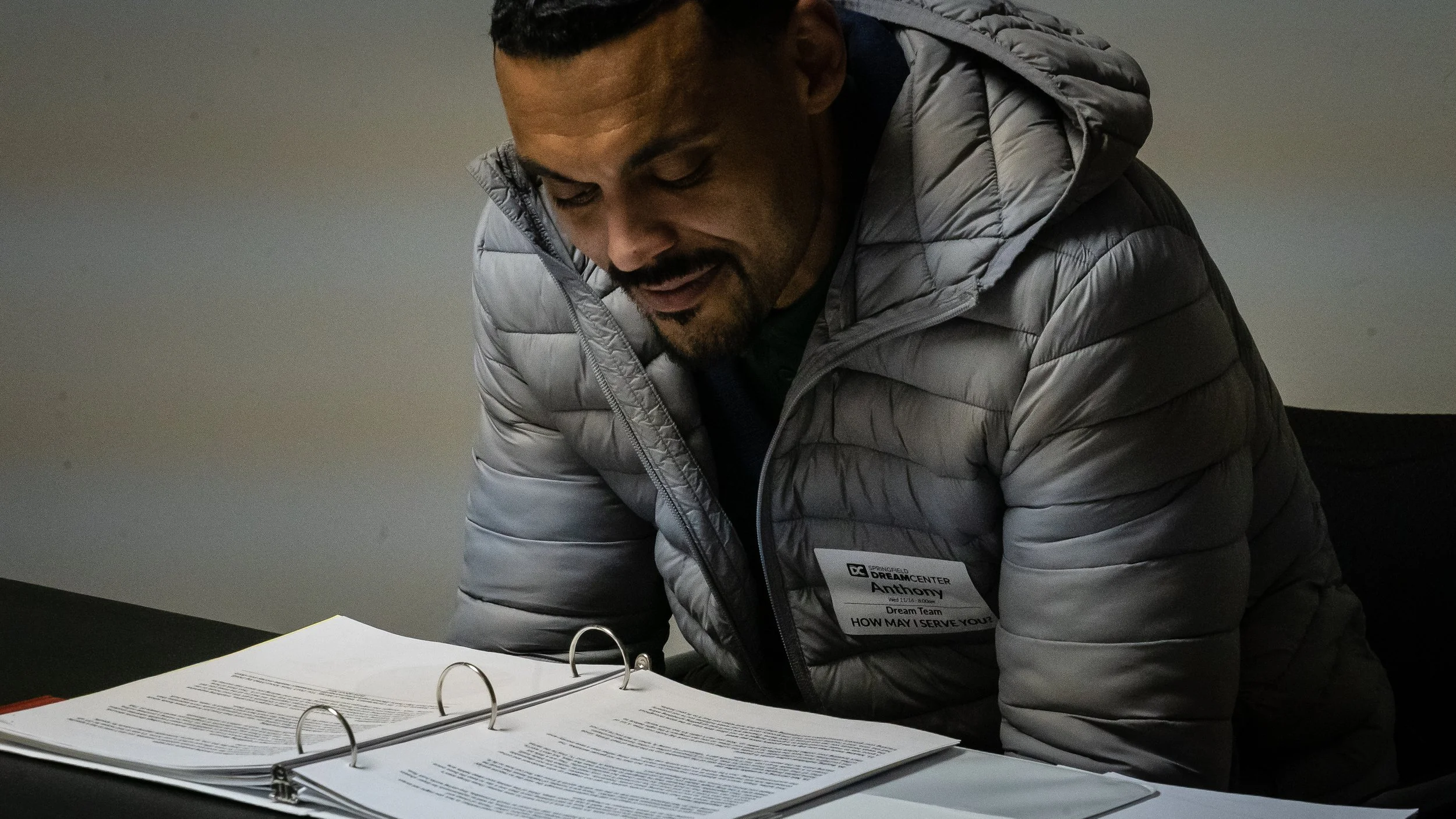 A man in a gray puffer jacket leaning over a table with open binder folders filled with papers, smiling while reading.