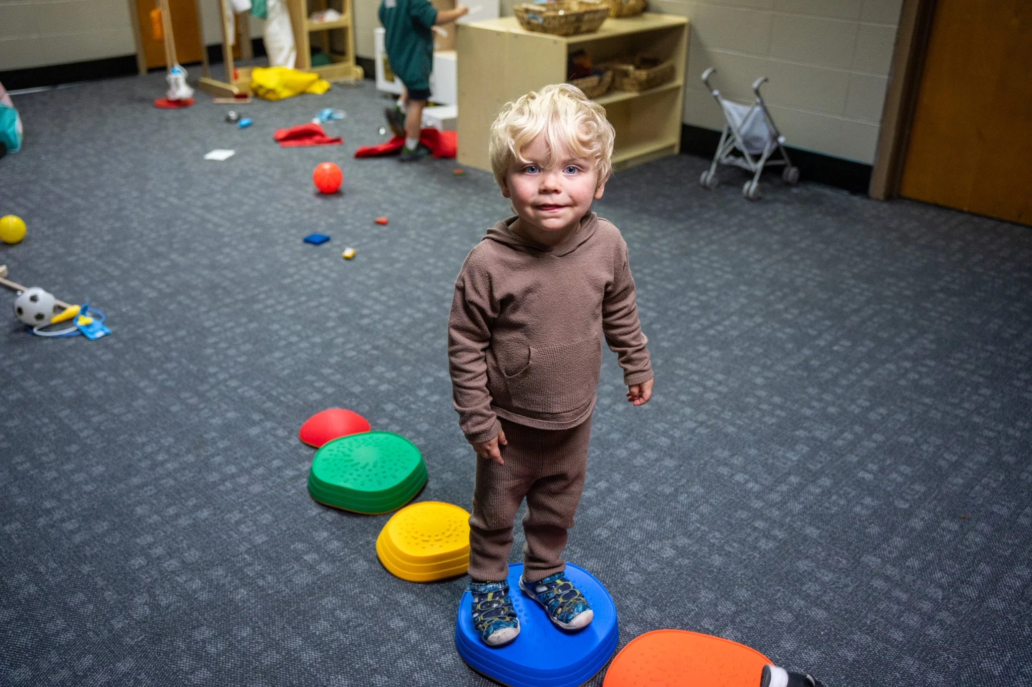 A young boy with blonde curly hair and a brown hoodie and pants stands on a blue balance pod in a playroom. The room has various toys scattered on the carpeted floor, including a red ball, a yellow disc, an orange disc, and other play items. In the background, there are shelves, a stroller, and other children playing.