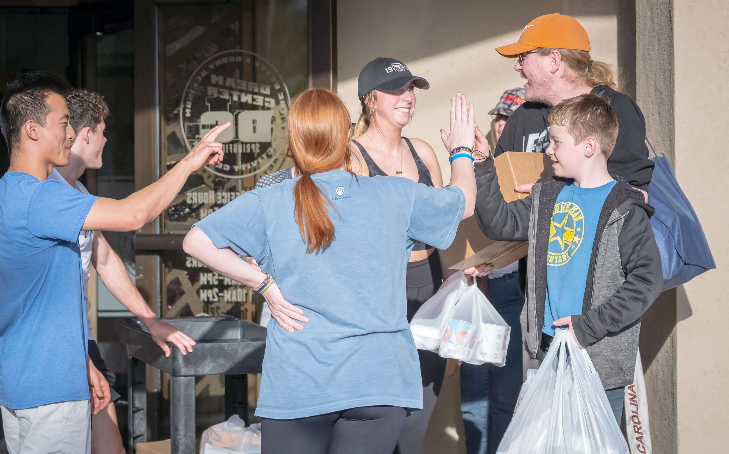 A group of young people and a woman exchanging high fives outside a building during a donation or charity event, with some holding grocery bags.
