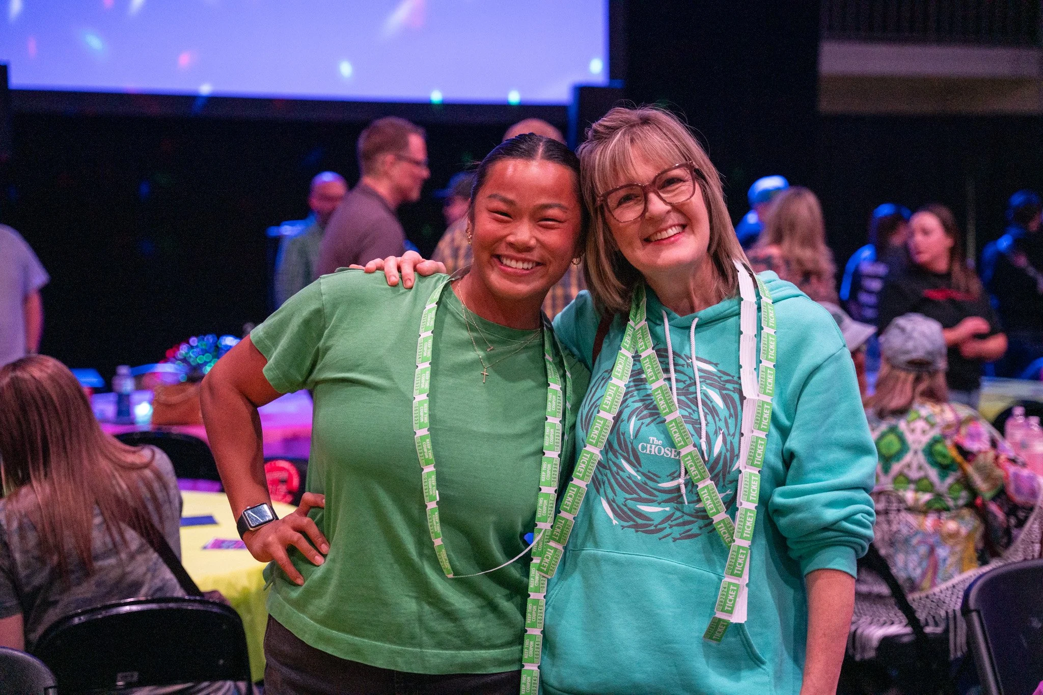 Two women smiling and posing together at an event, with tickets hanging around their necks, in a lively indoor setting with people in the background.