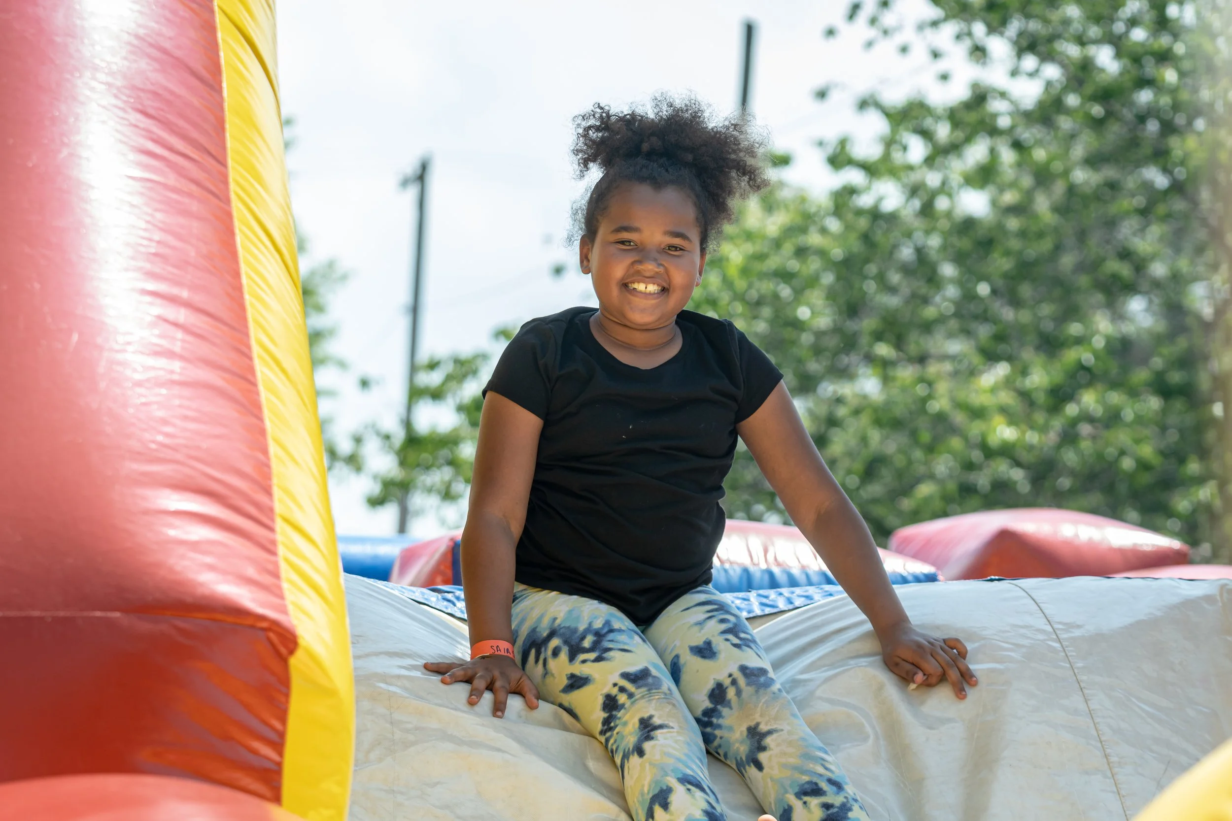 A young girl with curly hair smiling while sitting on an inflatable bounce house outdoors during daytime, with trees and utility poles in the background.