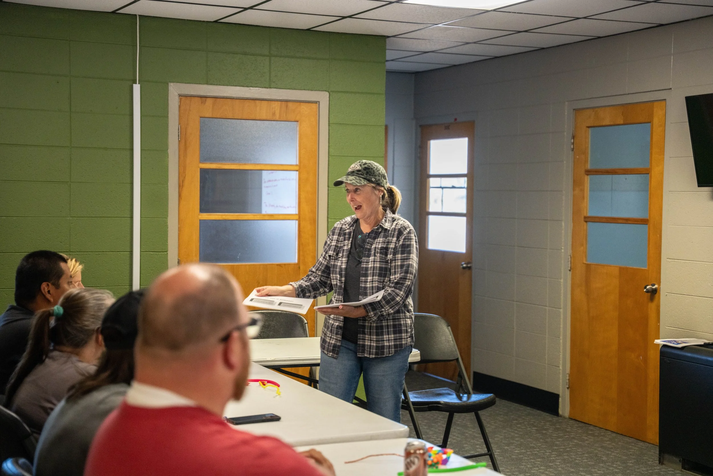 A woman in a plaid shirt and cap speaking to a group of people seated at tables in a room with green and gray walls and wooden doors.