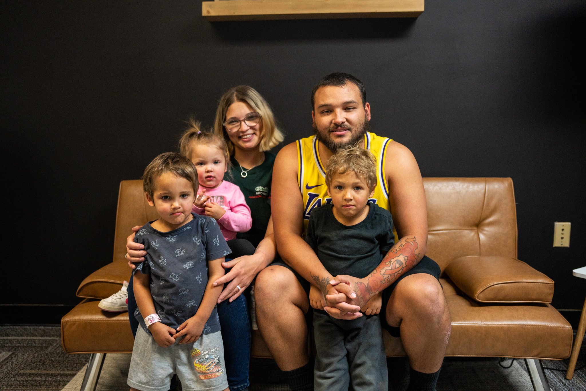 A family of five sitting on a brown leather sofa in a dimly lit room with a black wall. The father is wearing a yellow Los Angeles Lakers basketball jersey, and the mother is smiling with three children around them. The children are diverse in age, w