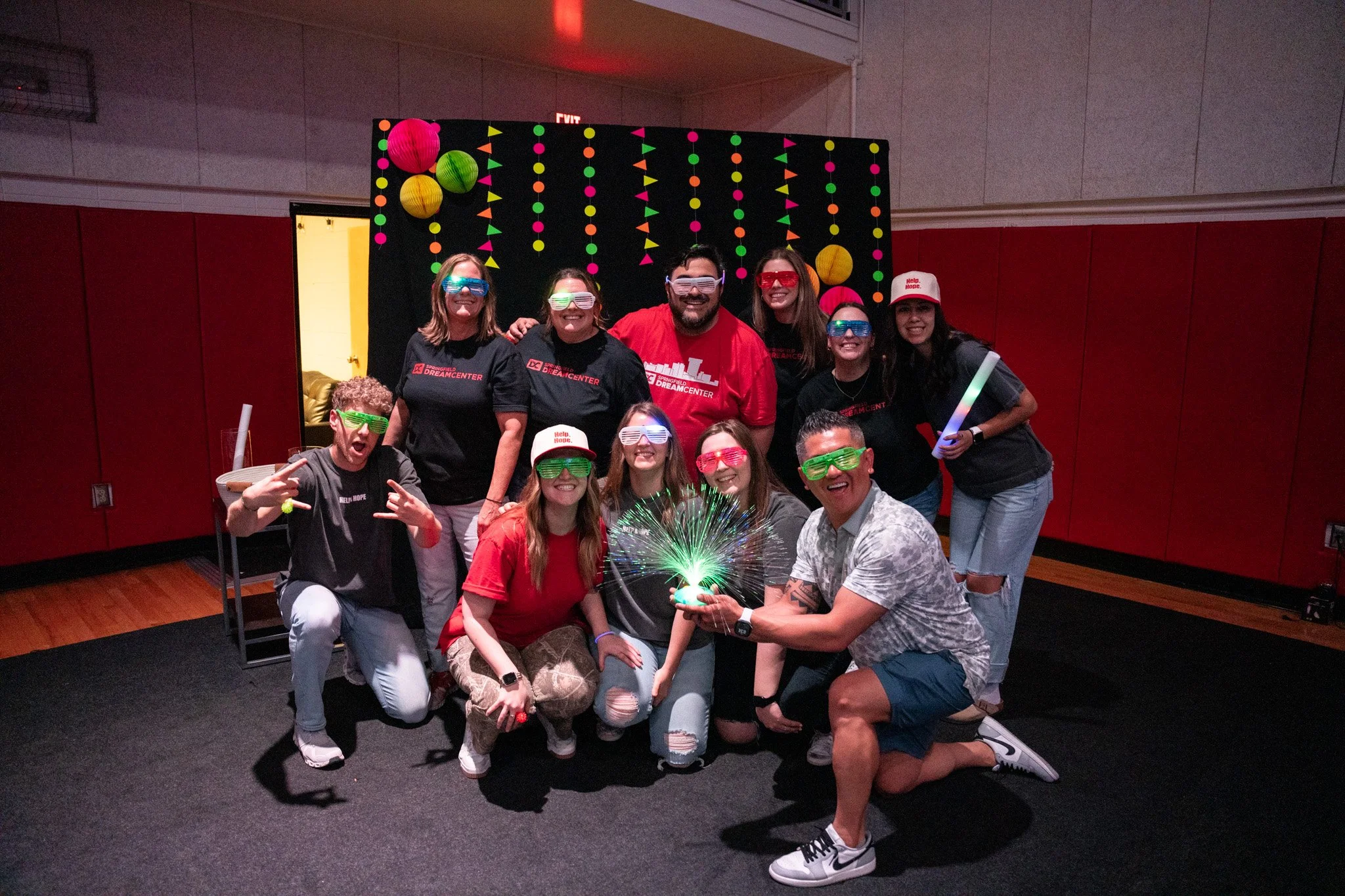 Group of people celebrating at a party, wearing colorful glow-in-the-dark glasses and holding a sparkler, with a decorated backdrop featuring colorful lanterns and paper flags.