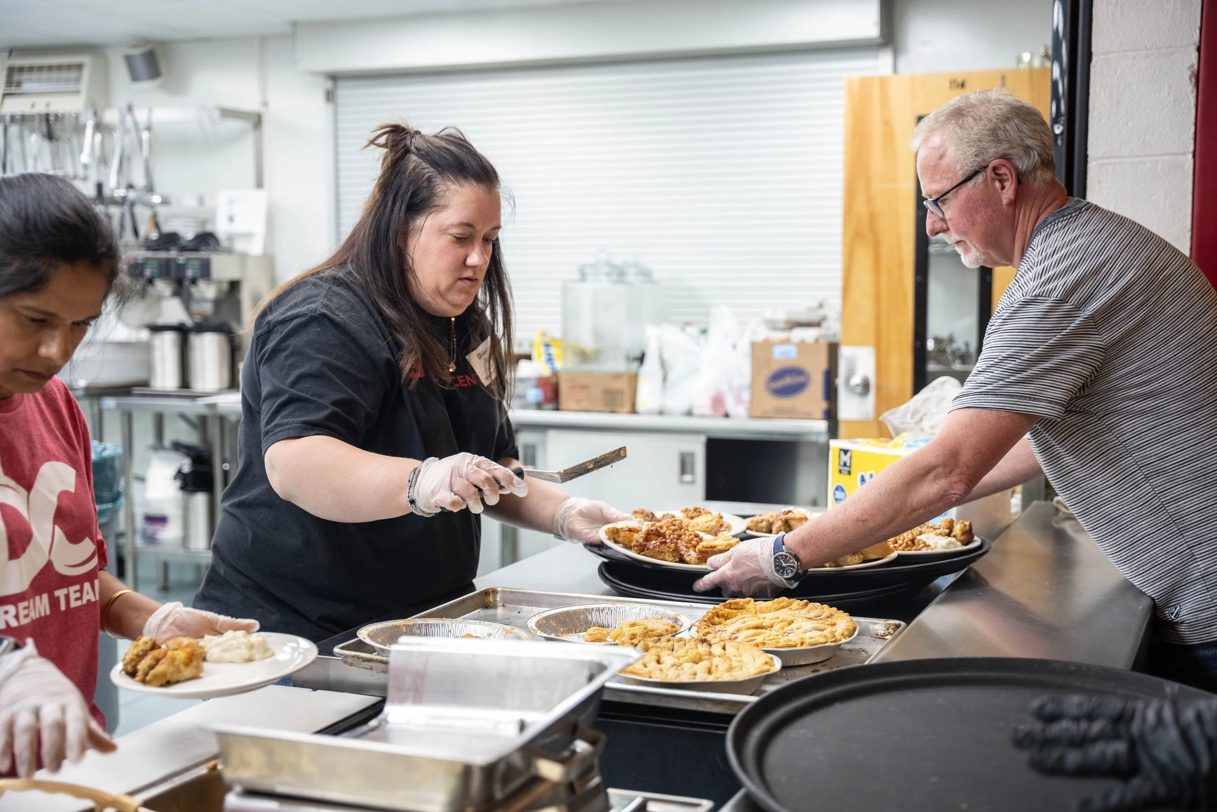 People serving food in a commercial kitchen, filling plates with fried chicken and pie.